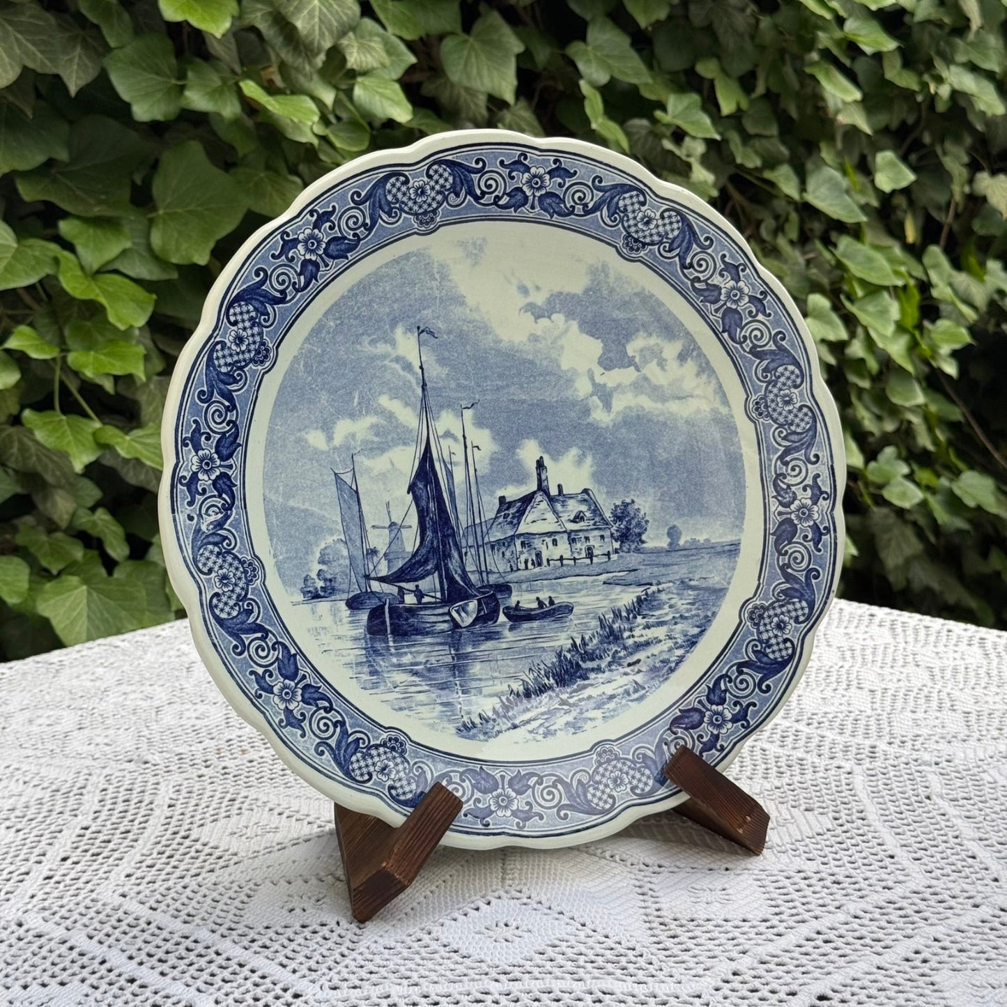 Decorative plate with a blue and white scene of a boat and house on a stand against a green leafy background.
