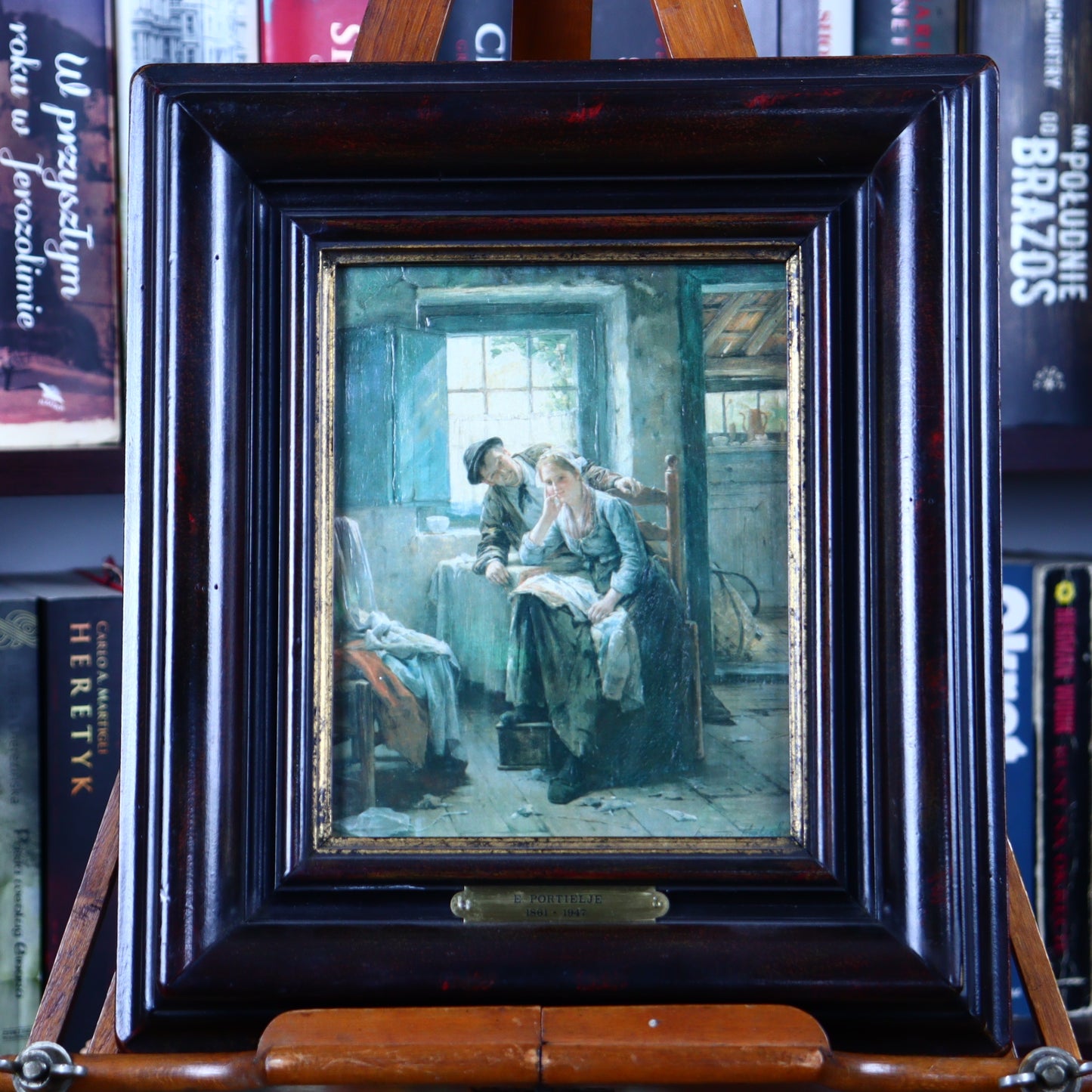 Framed artwork depicting a scene with a woman reading to a child in a room with a window and a chair. The frame is made of wood and the artwork is in oil paint.