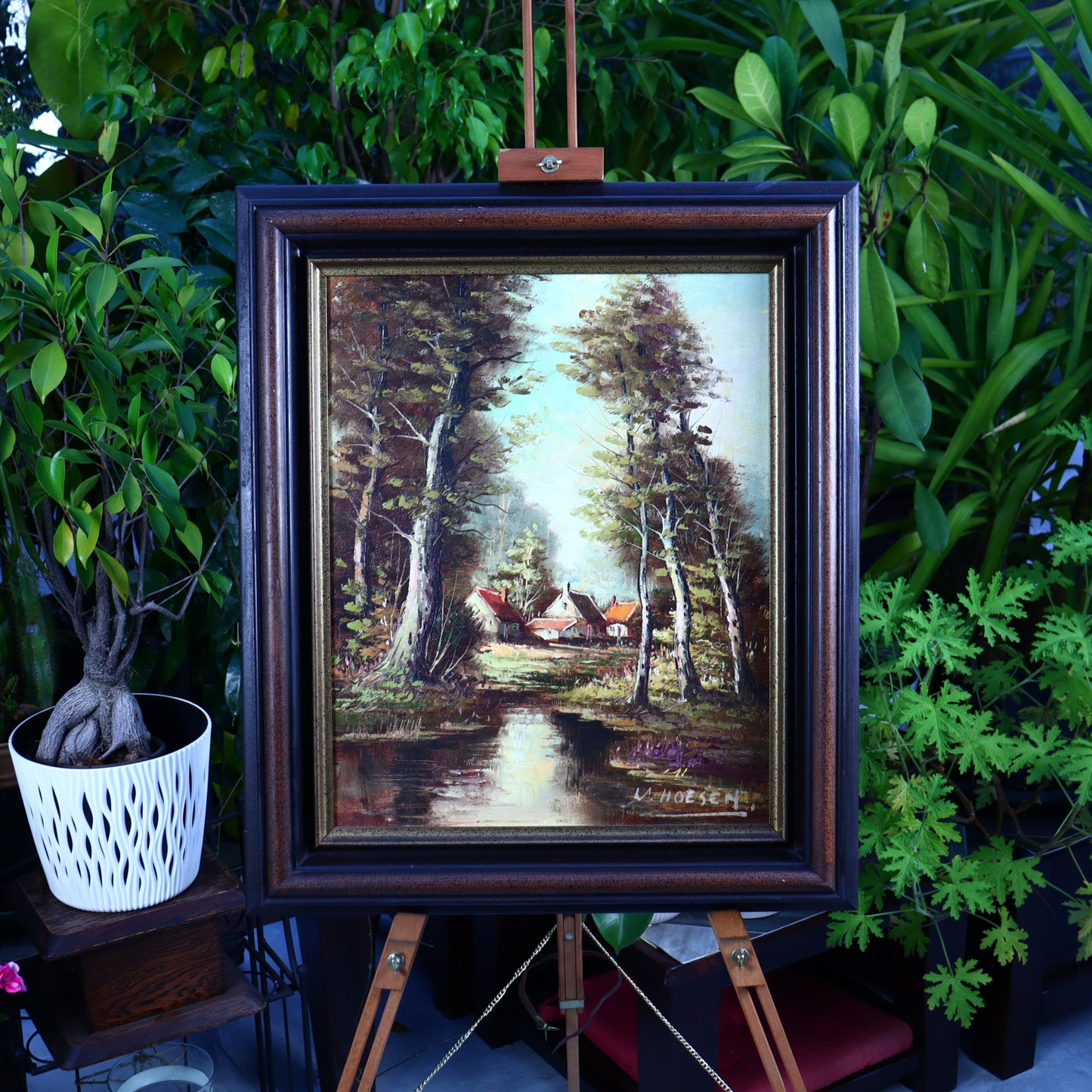 A framed oil painting on canvas depicting a landscape with cottages and trees, signed by W. Hoessen, displayed in a garden setting.