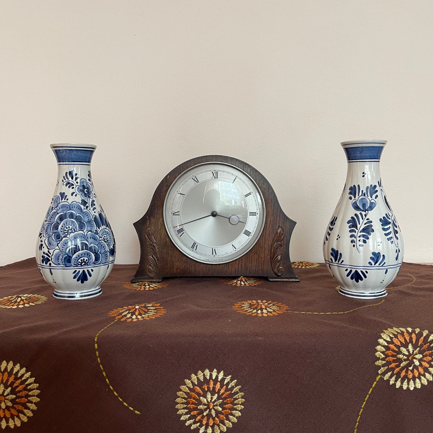 Wooden clock with two blue and white floral vases on a tablecloth with floral patterns.