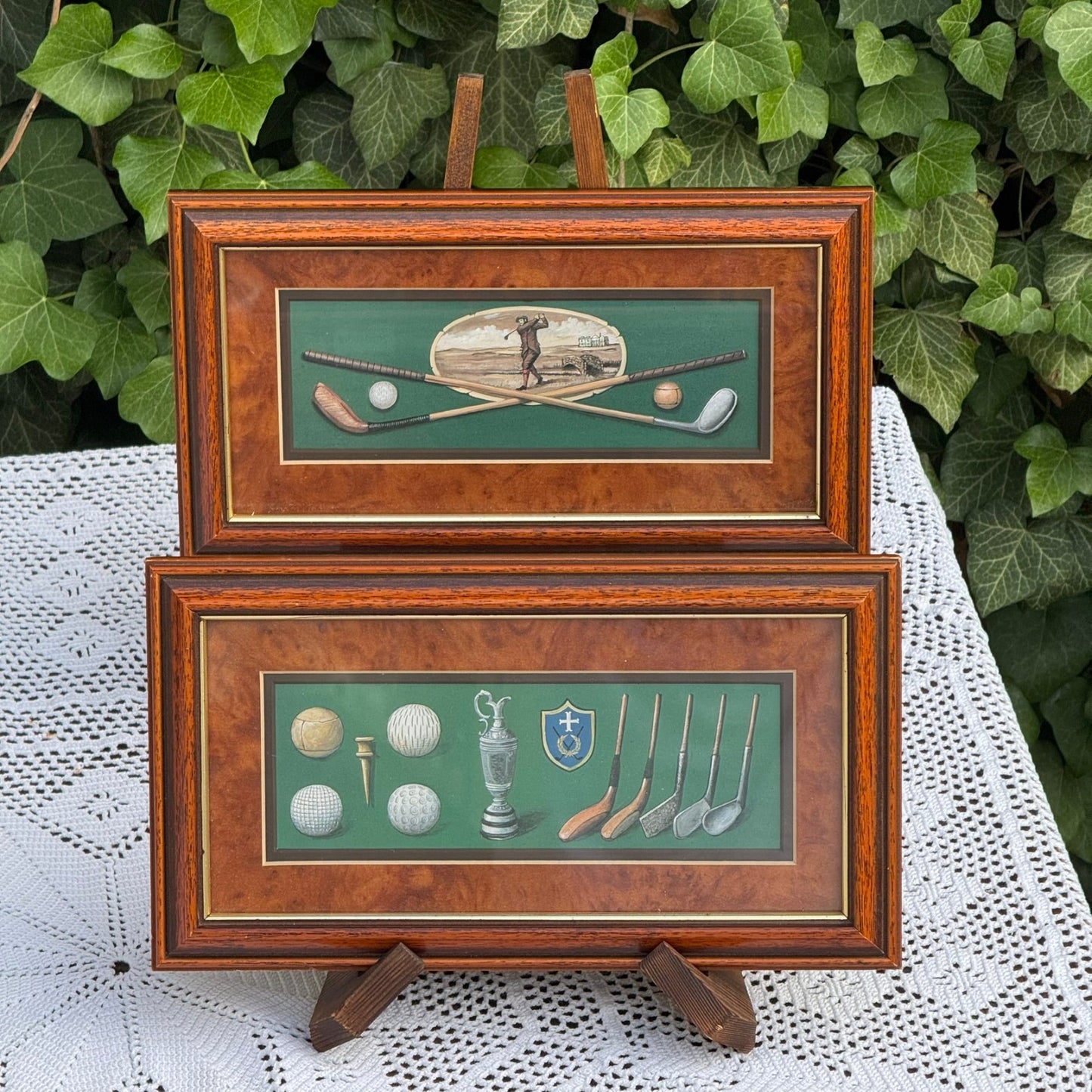 Two framed pictures of golf-themed artwork on a white lace tablecloth with green foliage in the background.