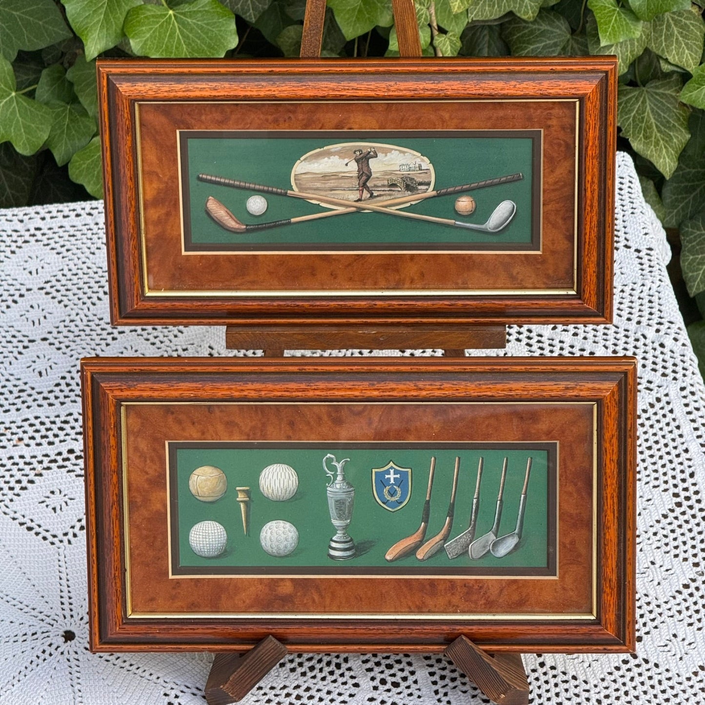 Two framed pictures of golf-themed artwork on a lace tablecloth with green foliage in the background.