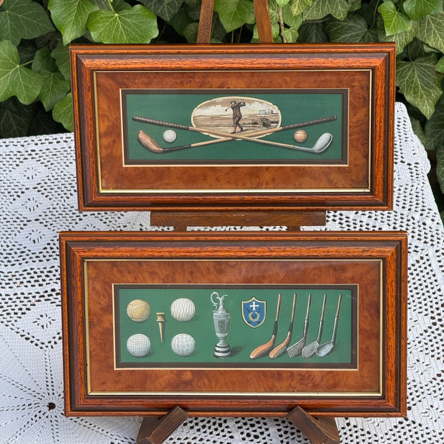 Two framed pictures of golf-themed artwork on a white lace tablecloth with green foliage in the background.