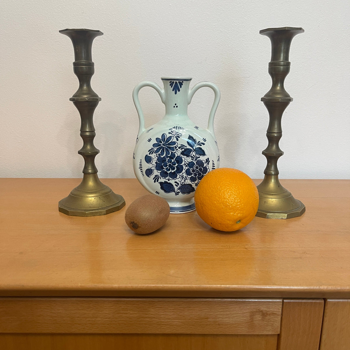 Decorative items including a white vase with blue floral patterns, an orange, a kiwi, and two brass candlesticks on a wooden surface.