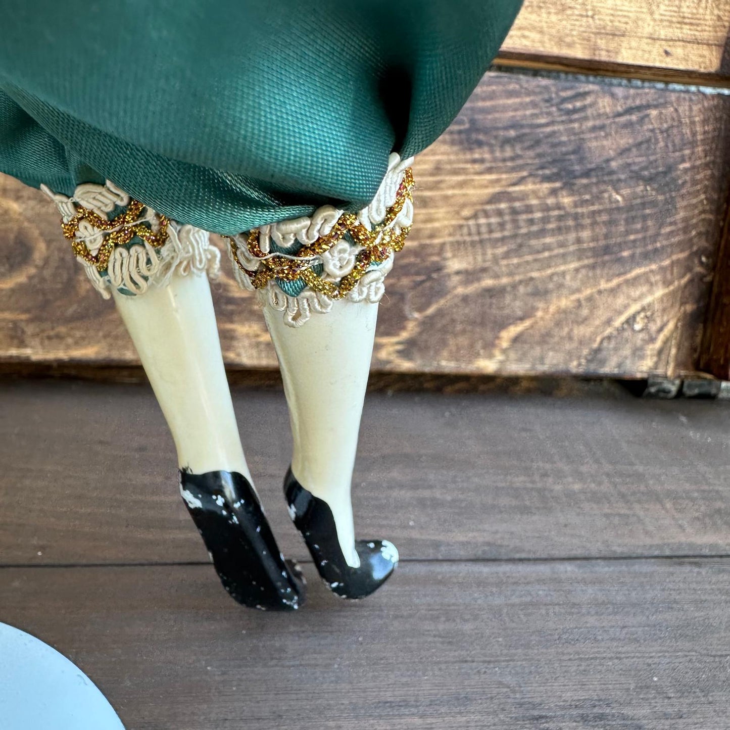 Close-up of a person wearing green pants with decorative white and gold garters on a wooden floor.