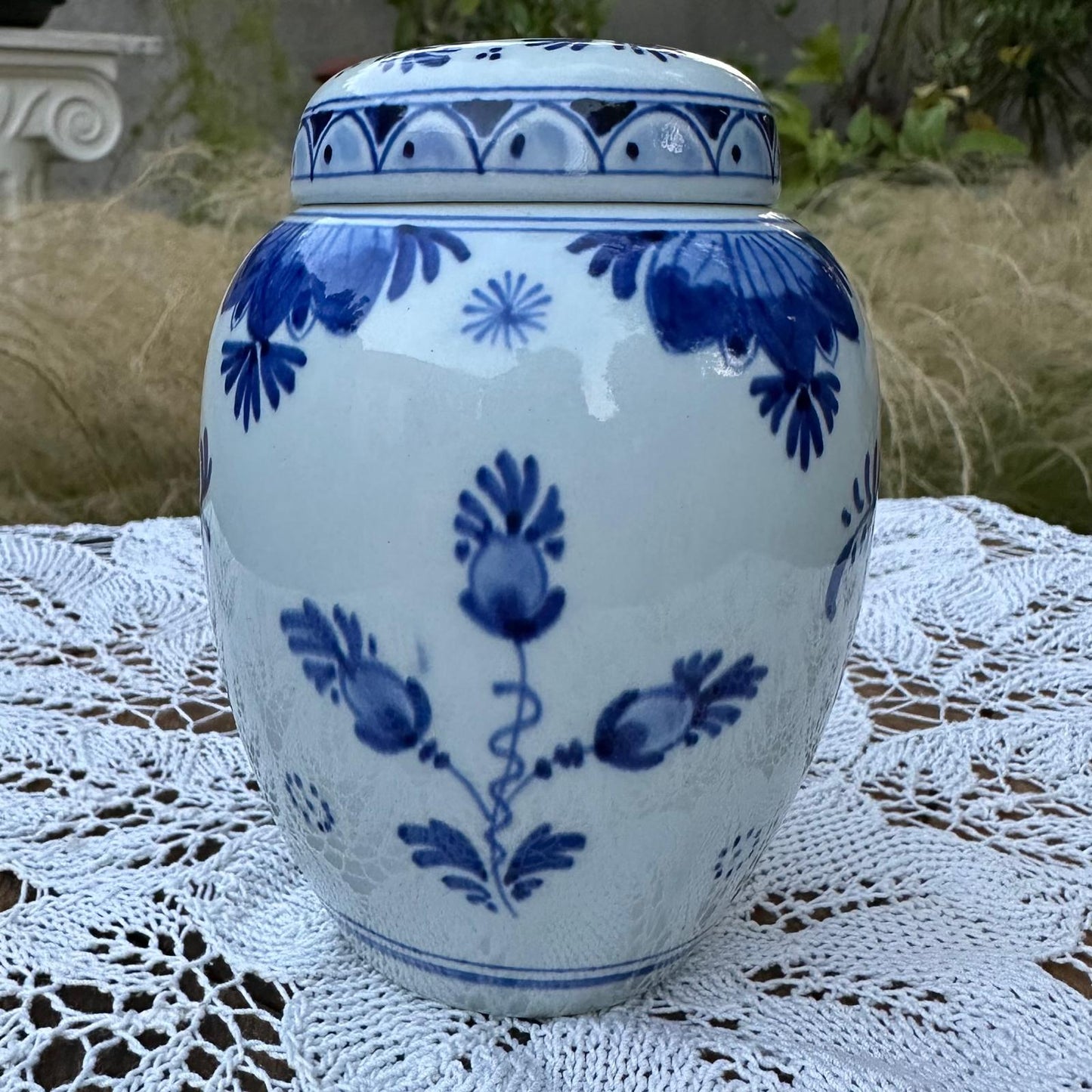 Blue and white ceramic jar with floral patterns on a lace tablecloth outdoors.
