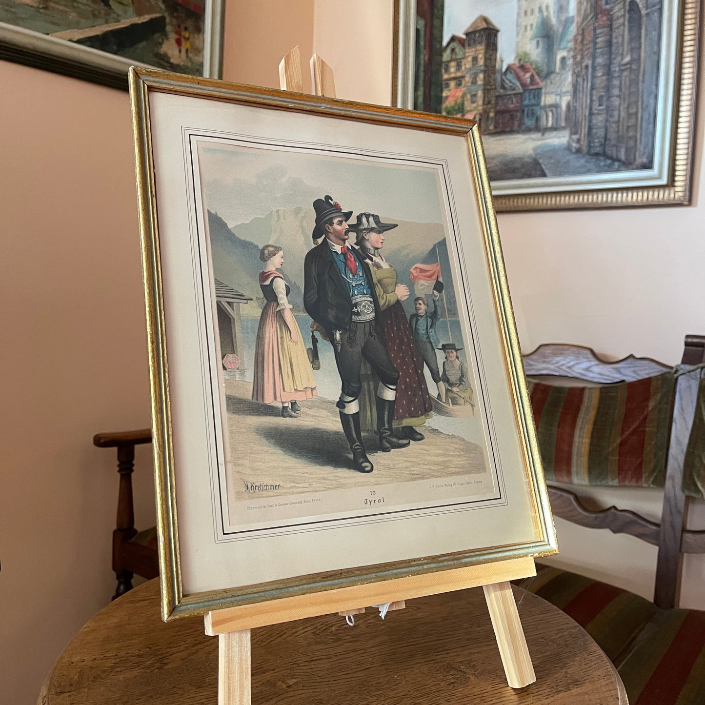 Framed artwork of a historical scene with people on an easel in a room with other framed pictures.