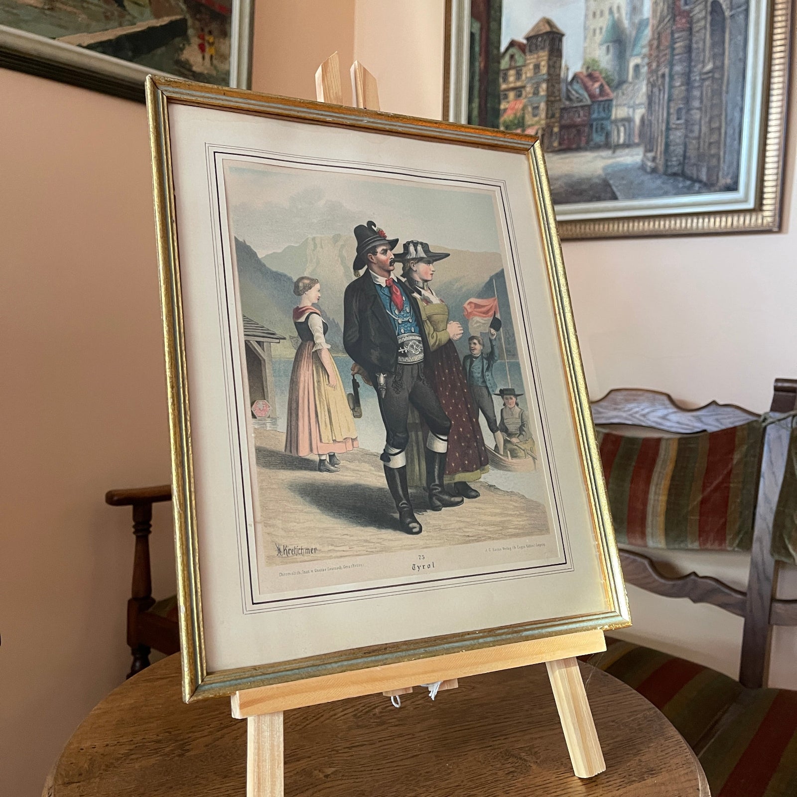 Framed artwork of a historical scene with people on an easel in a room with other framed pictures.