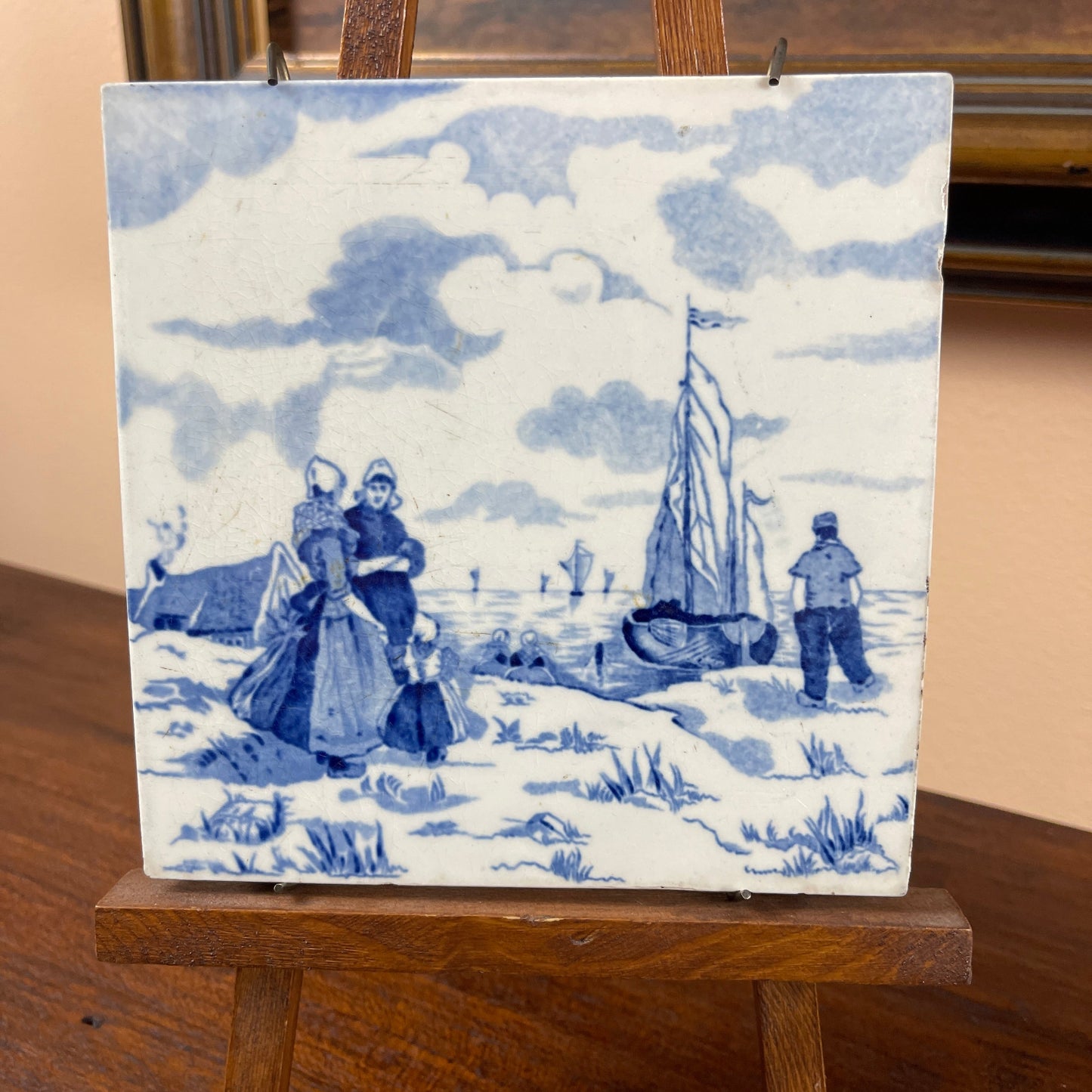 Decorative tile with blue and white design of people and a sailboat on a wooden stand.
