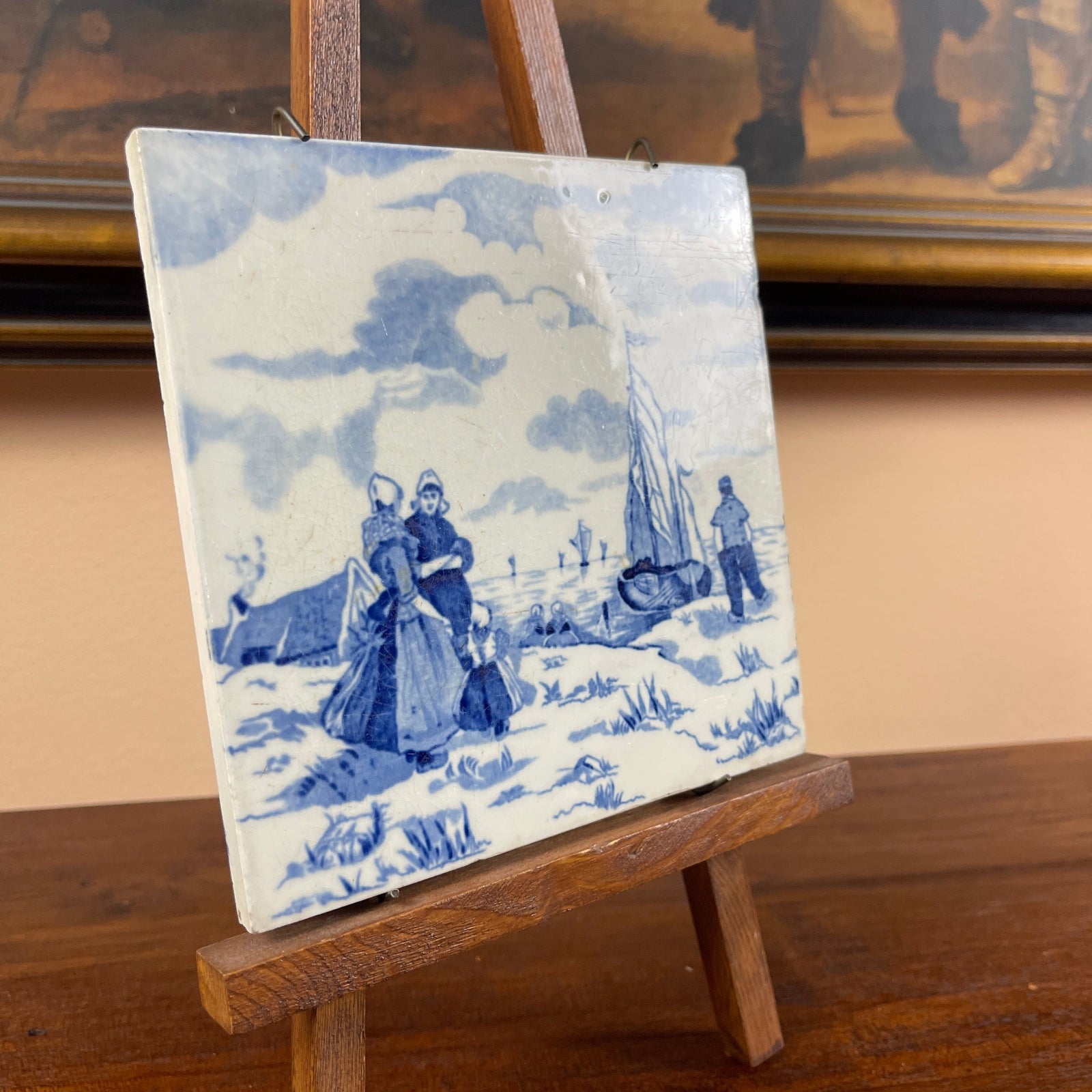 Decorative tile with blue and white design of people in a snowy landscape on a wooden easel.