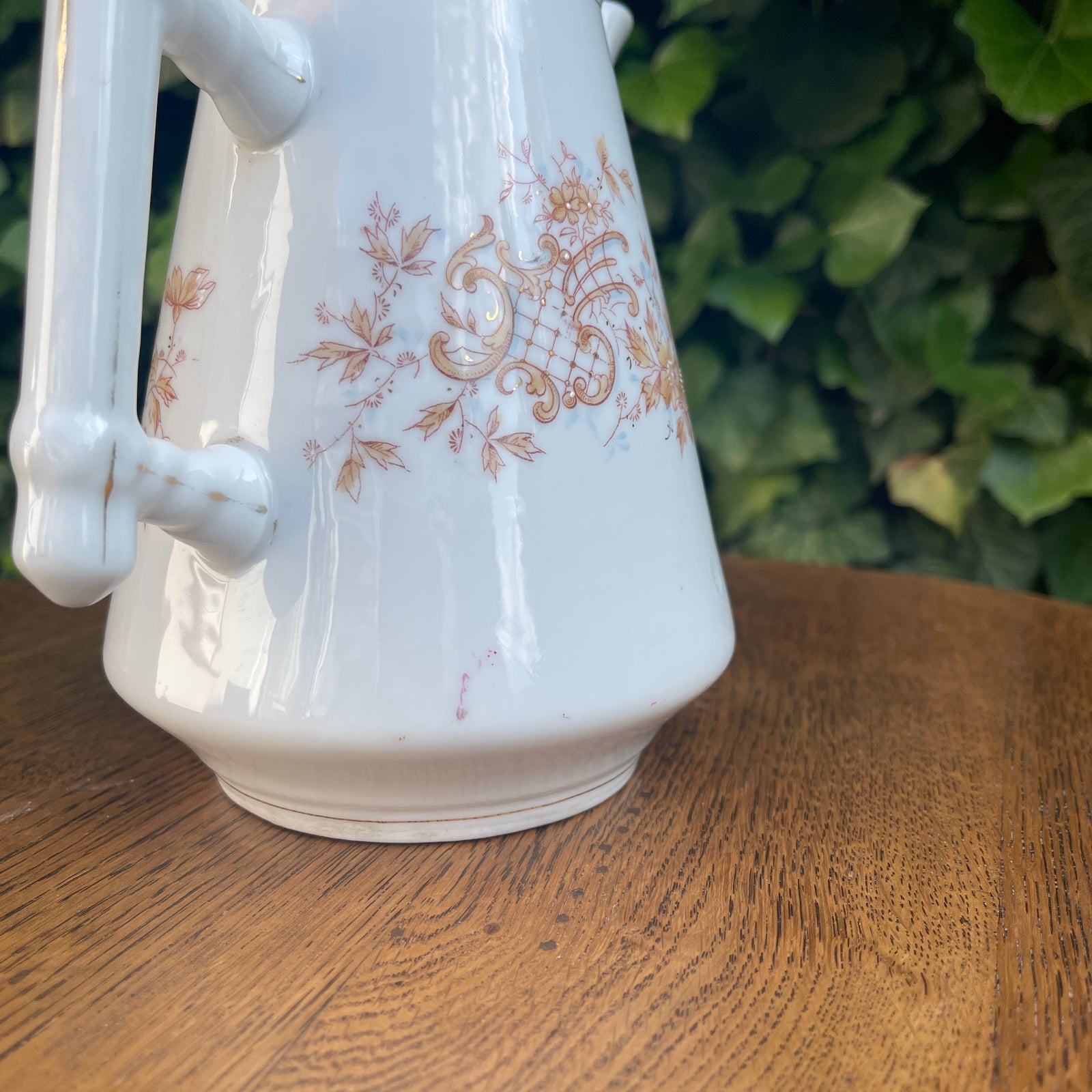 White ceramic teapot with gold floral patterns on a wooden surface with a green leafy background
