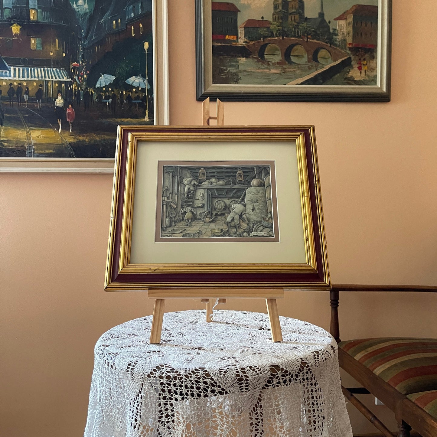 Framed artwork on a small table with lace tablecloth, surrounded by other framed pictures on a wall.