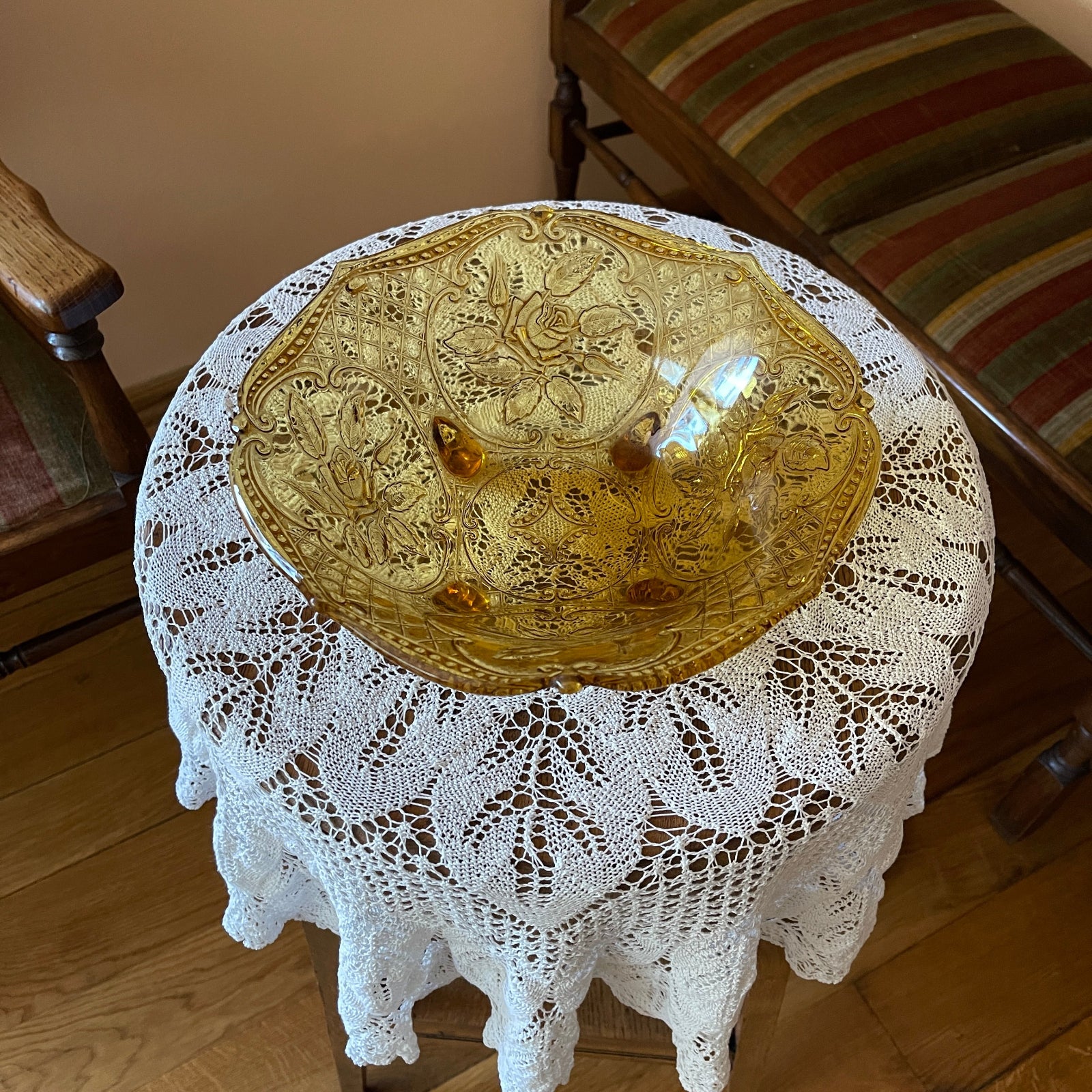 Decorative gold bowl on a round table with a lace tablecloth, surrounded by wooden chairs.