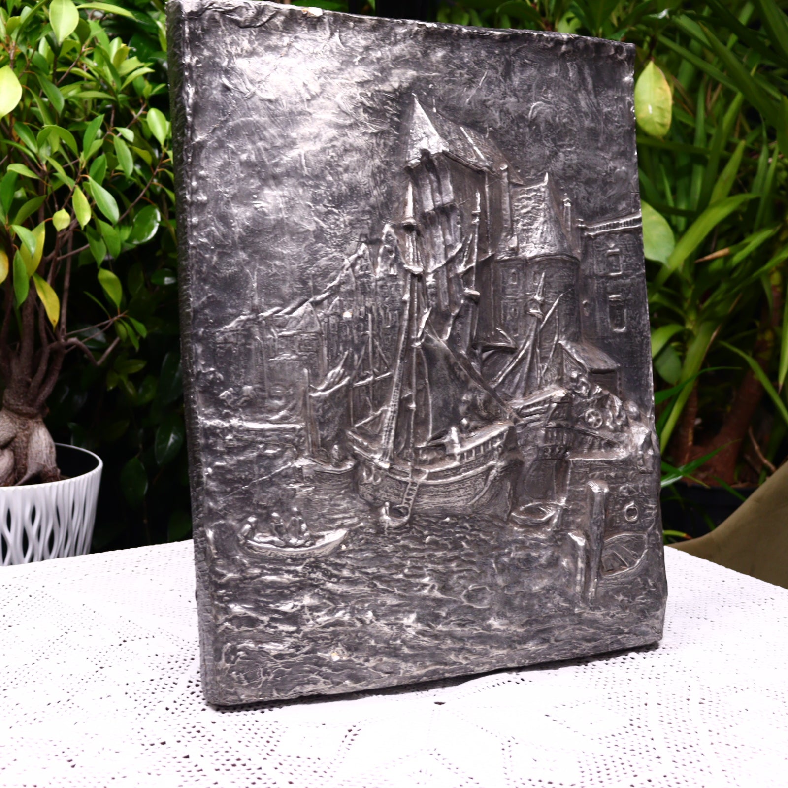 A concrete bas-relief depicting a medieval port city with an old crane, placed on a table with a plant background.