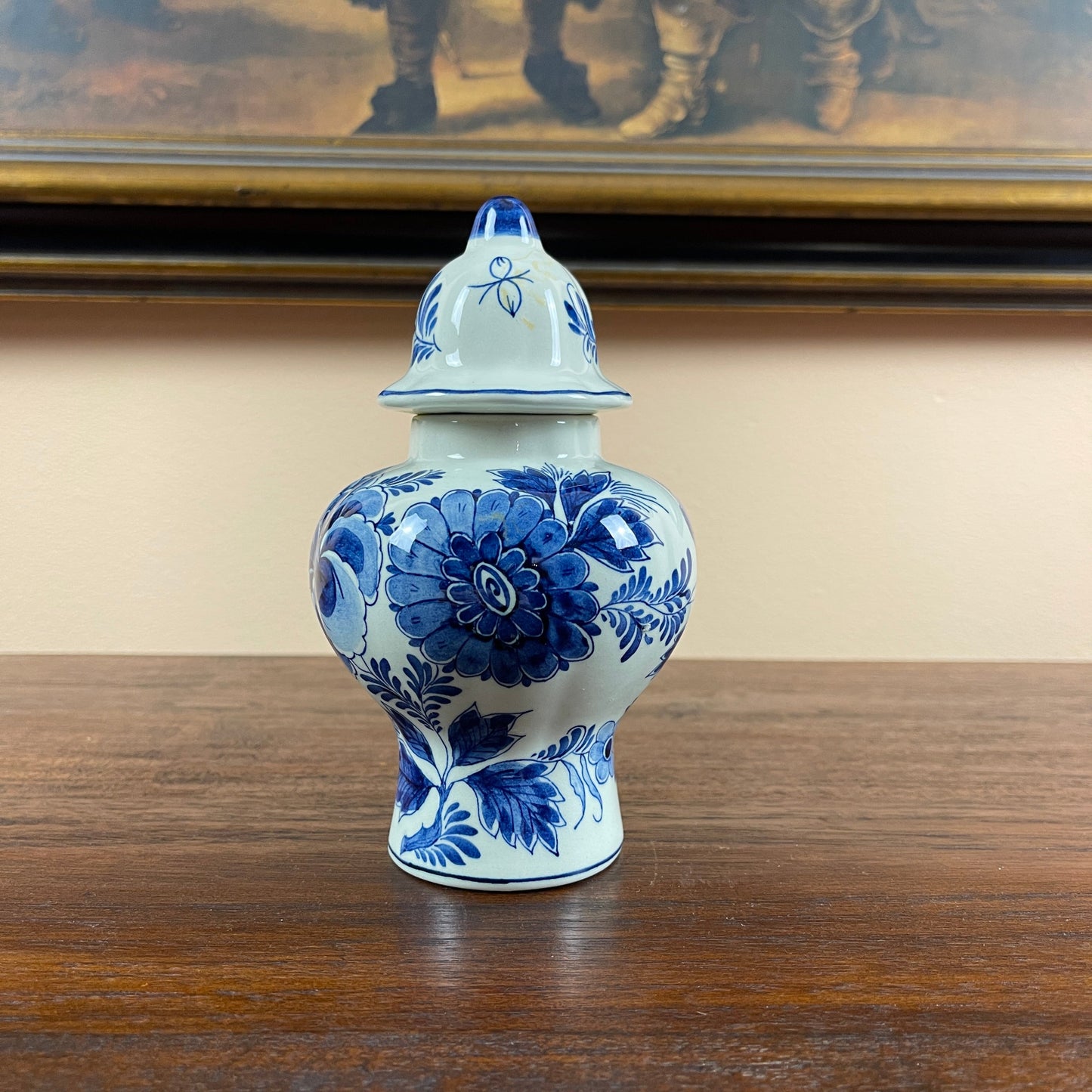 Blue and white floral-patterned ceramic jar on a wooden surface with a painting in the background.