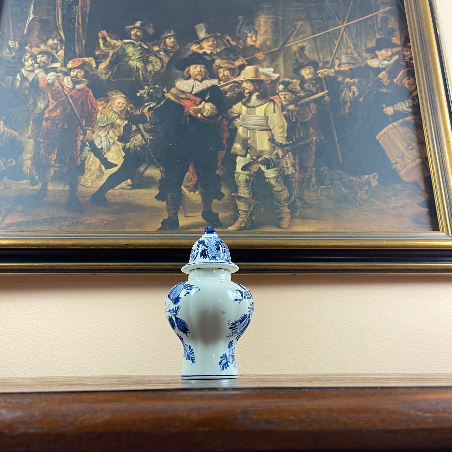 Blue and white ceramic vase on a table with a large painting of a historical scene in the background.