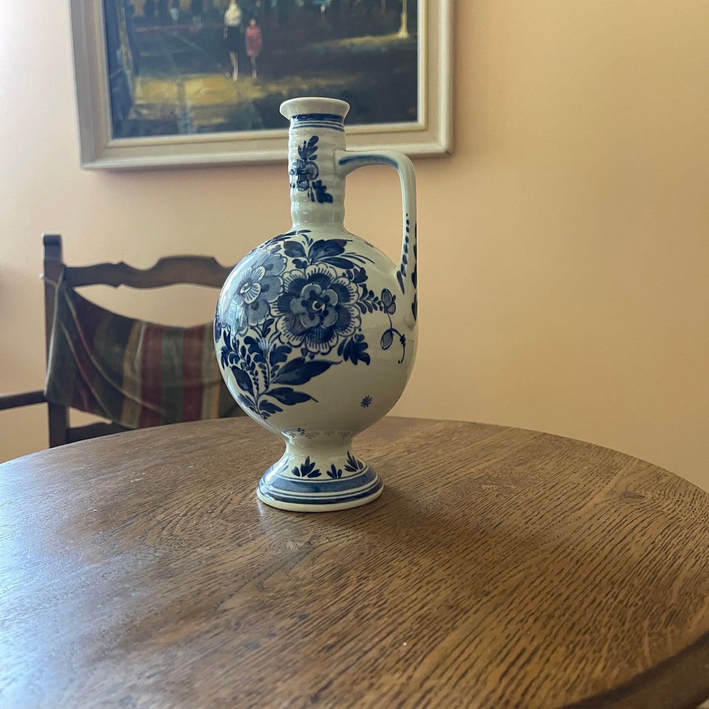 Blue and white floral ceramic vase on a wooden table with a chair and framed picture in the background.