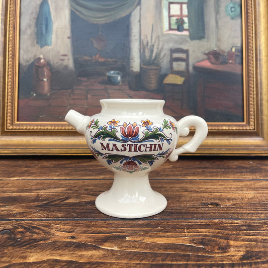 Decorative ceramic jar with 'Mastichin' text on a wooden surface in front of a framed painting.