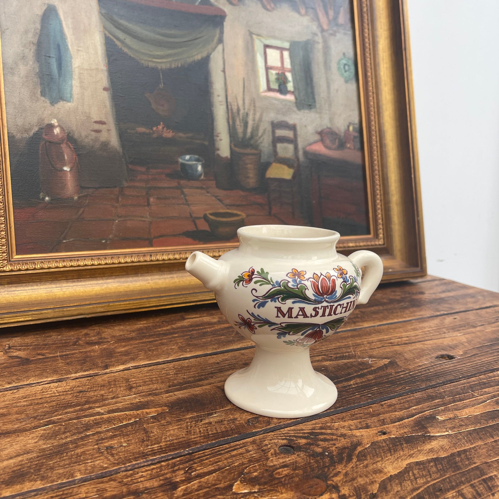 Decorative ceramic teapot with floral design on a wooden surface in front of a framed painting.