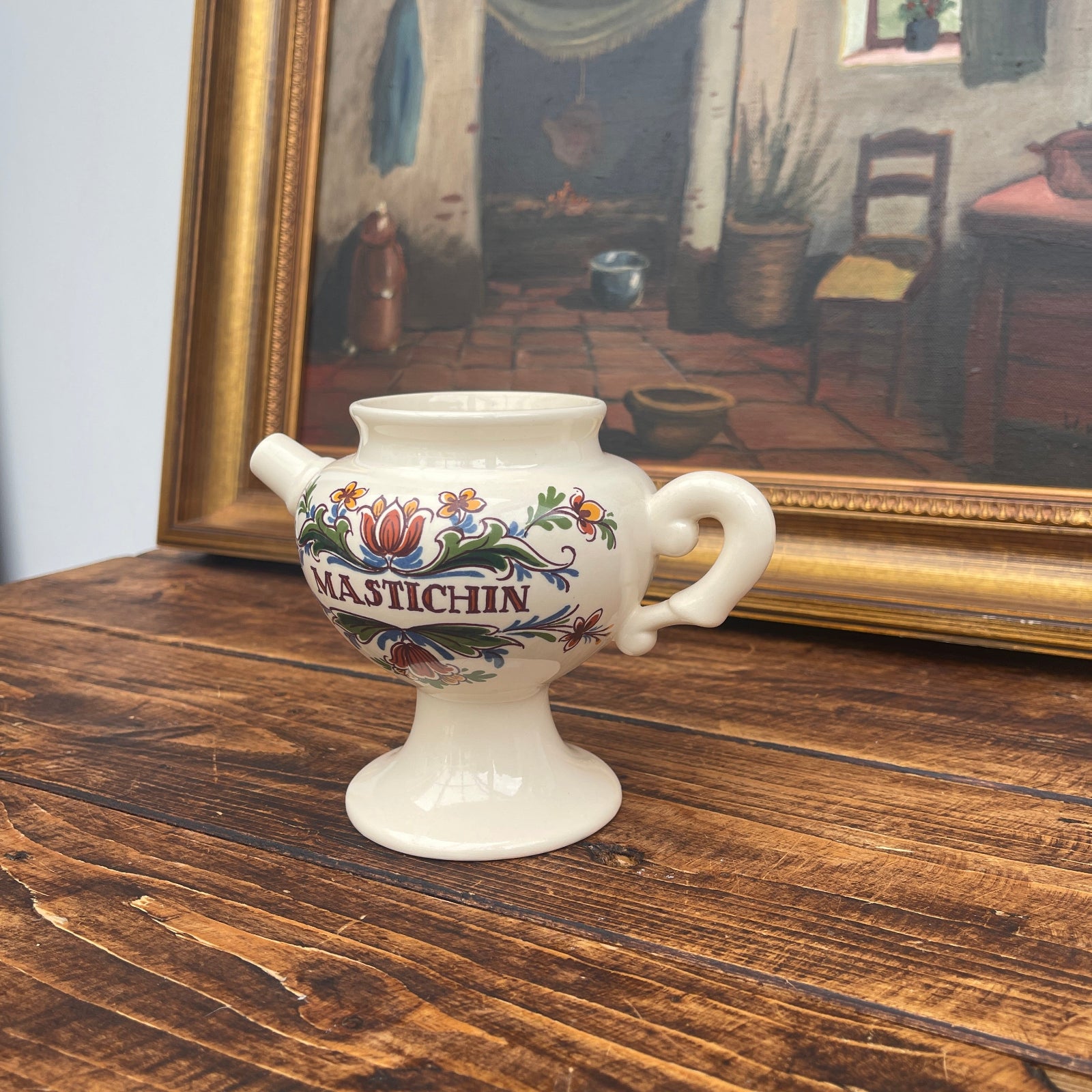 Decorative ceramic vase with floral designs and 'Mastichin' text on a wooden surface, with a painting in the background.