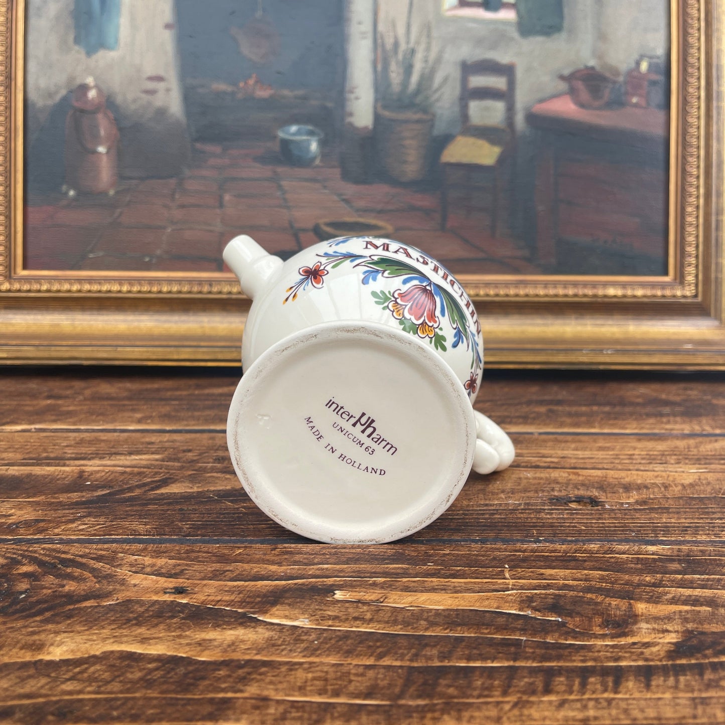 Decorative ceramic teapot with floral patterns on a wooden surface, framed artwork in the background.