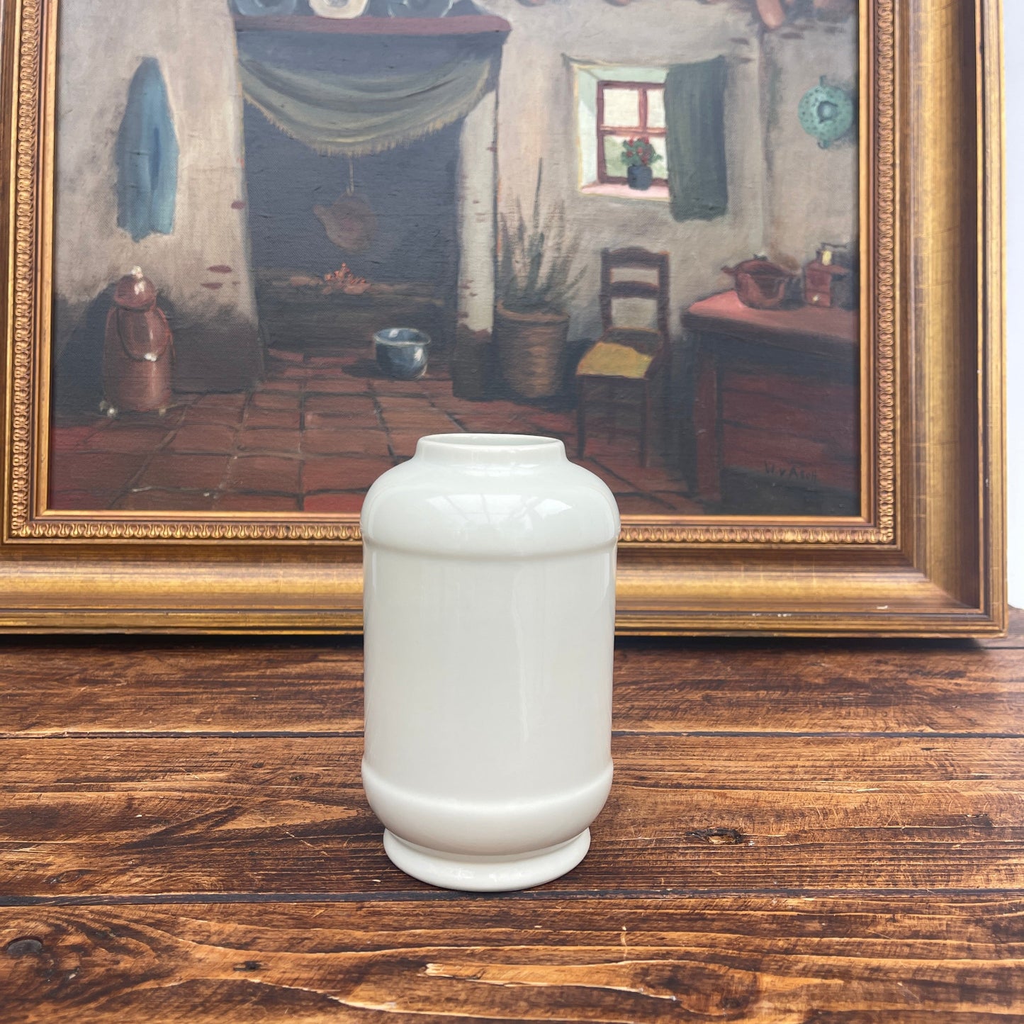 White ceramic vase on a wooden surface with a framed painting in the background