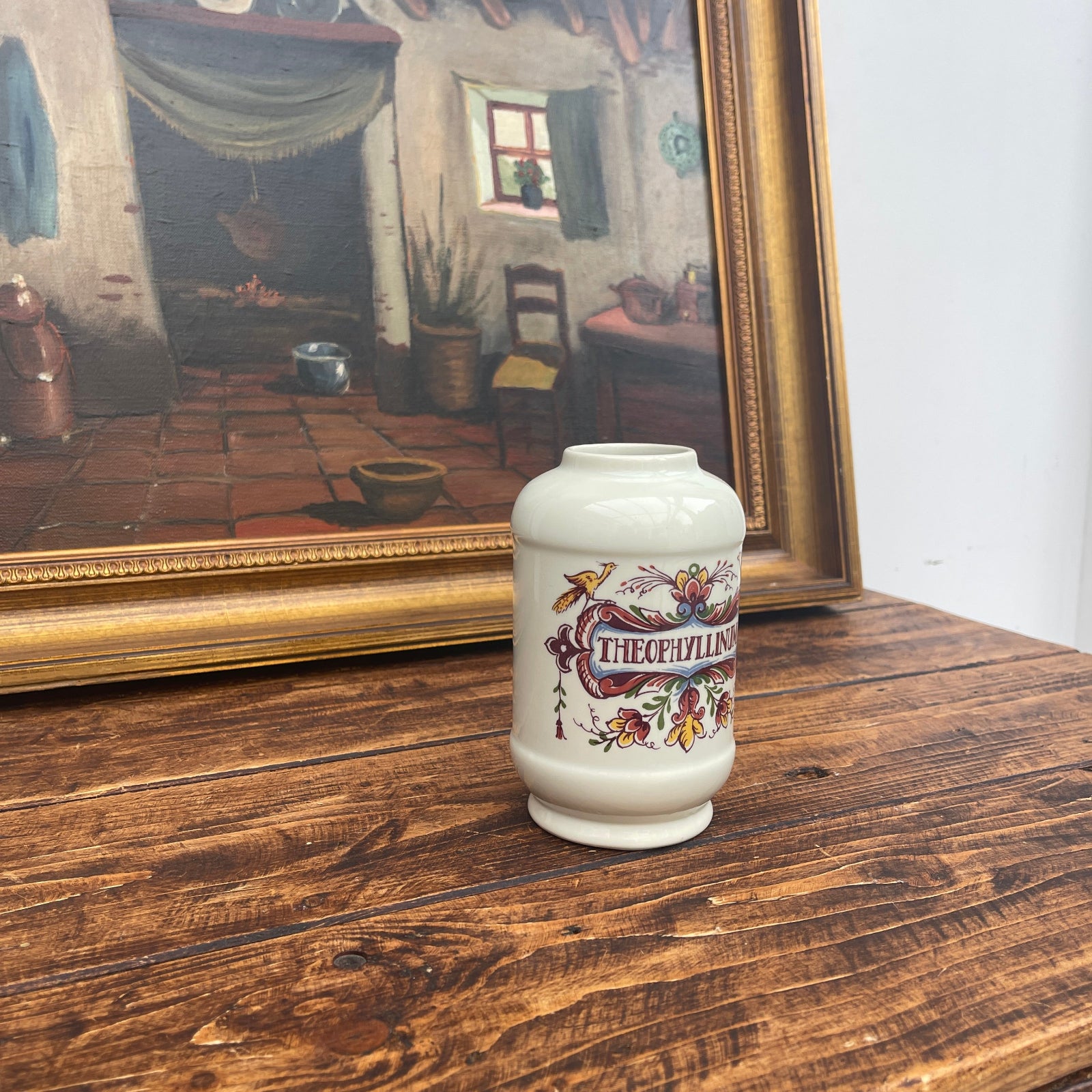 Decorative vase with 'Theophylac' design on a wooden surface in front of a framed painting.