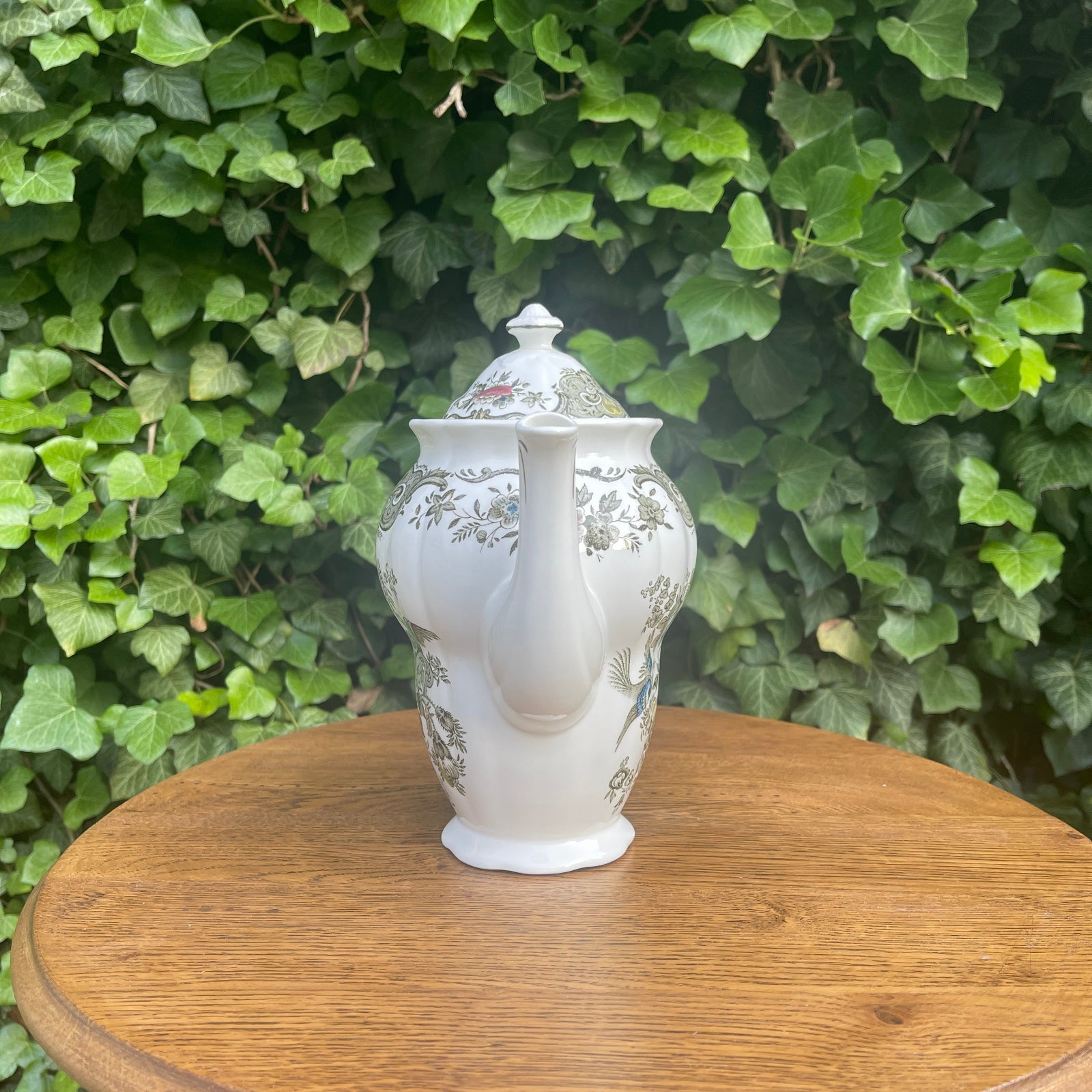 White ceramic teapot with floral patterns on a wooden surface with a green leafy background