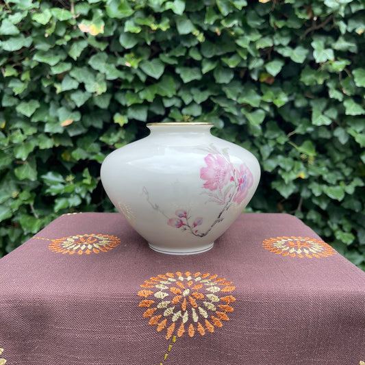 Decorative vase with floral design on a brown fabric surface with embroidered patterns, against a green leafy background.