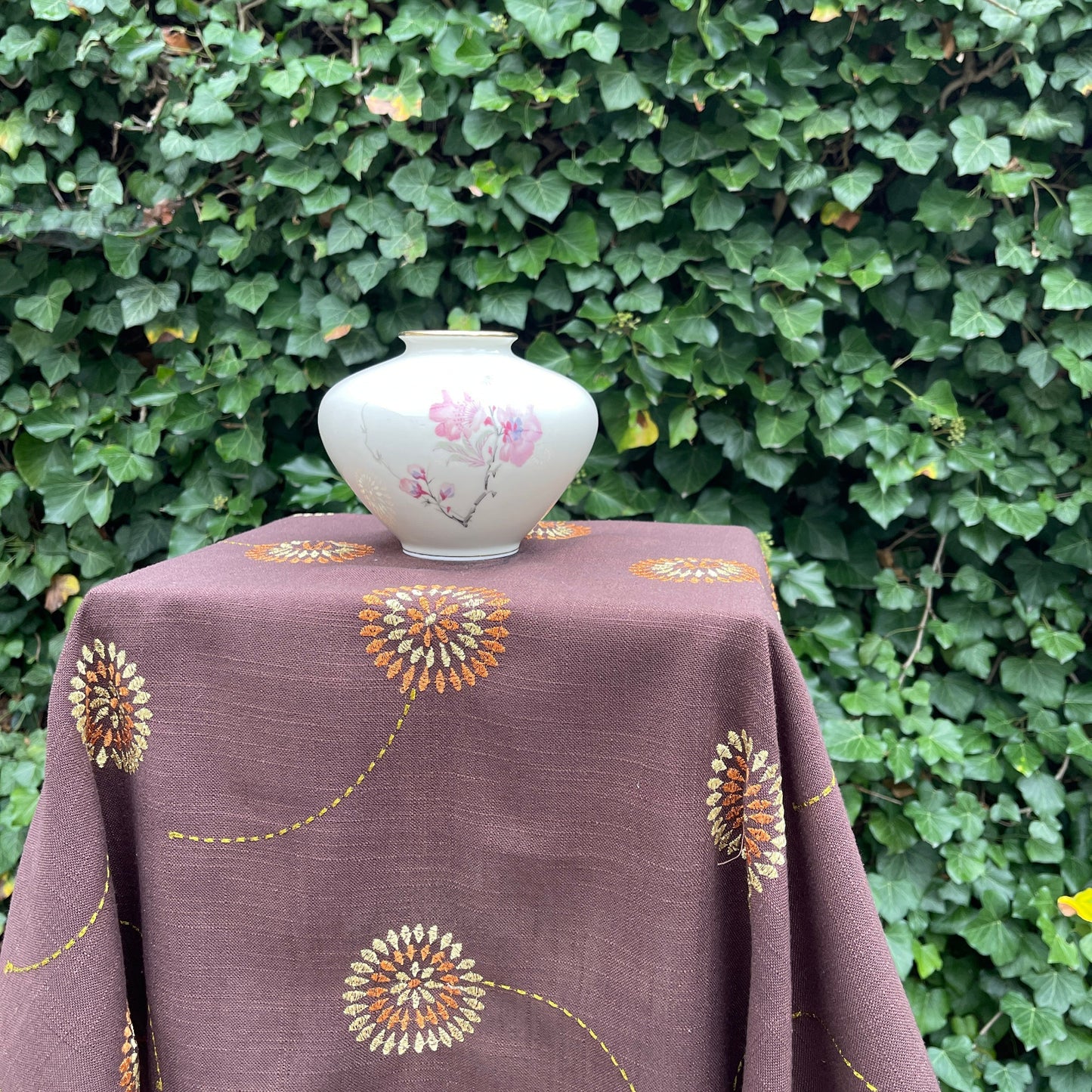 Vase on a tablecloth with floral design against a green hedge background