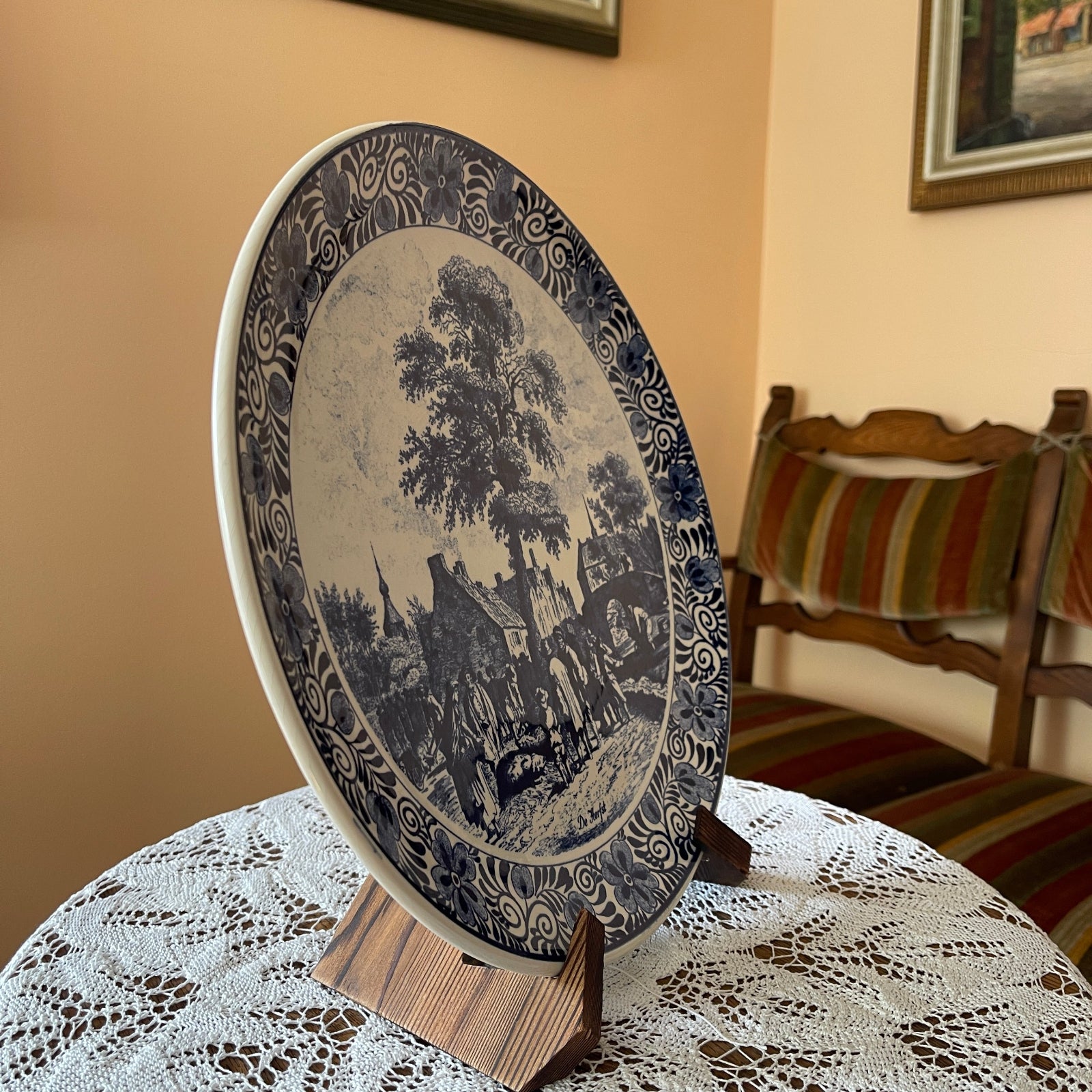 Decorative plate with landscape design on a stand in a room with a striped chair and lace tablecloth.