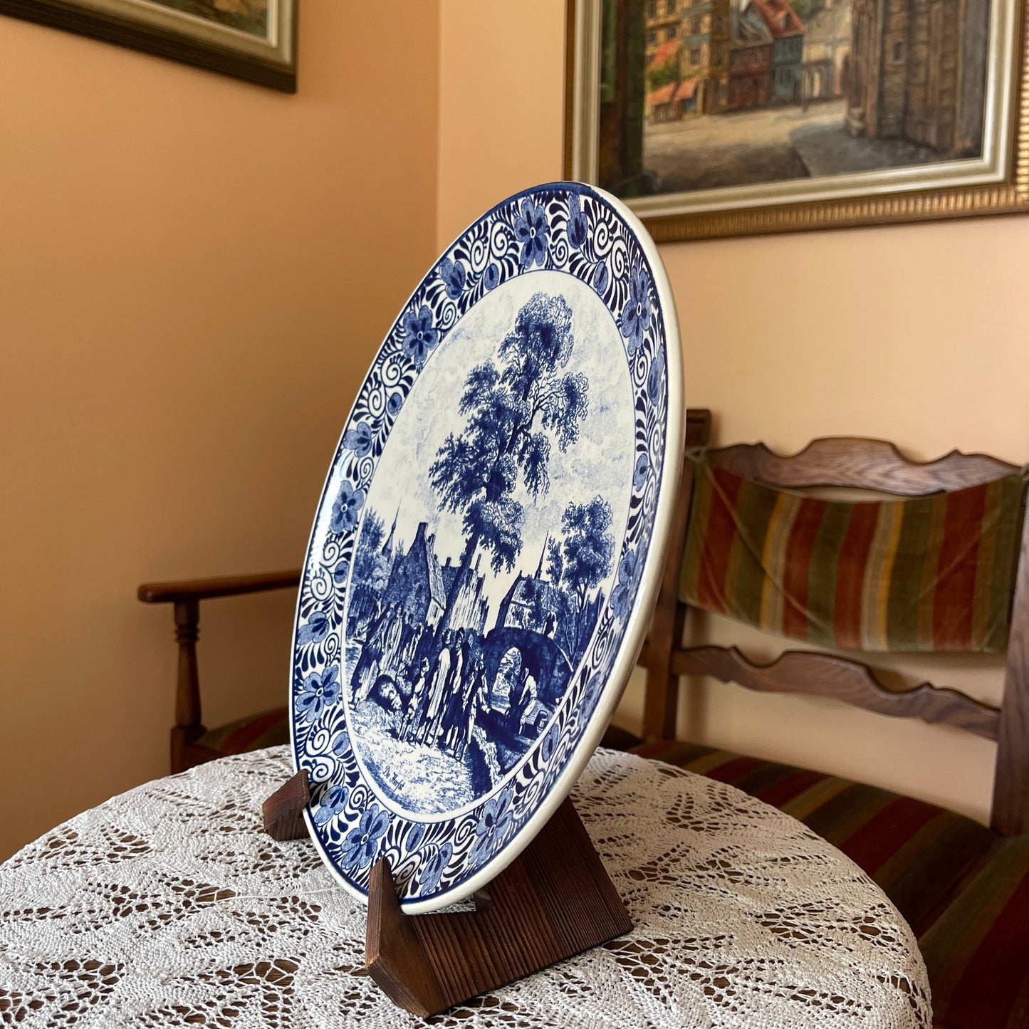 Blue and white decorative plate on a stand in a room with lace tablecloth and framed pictures.