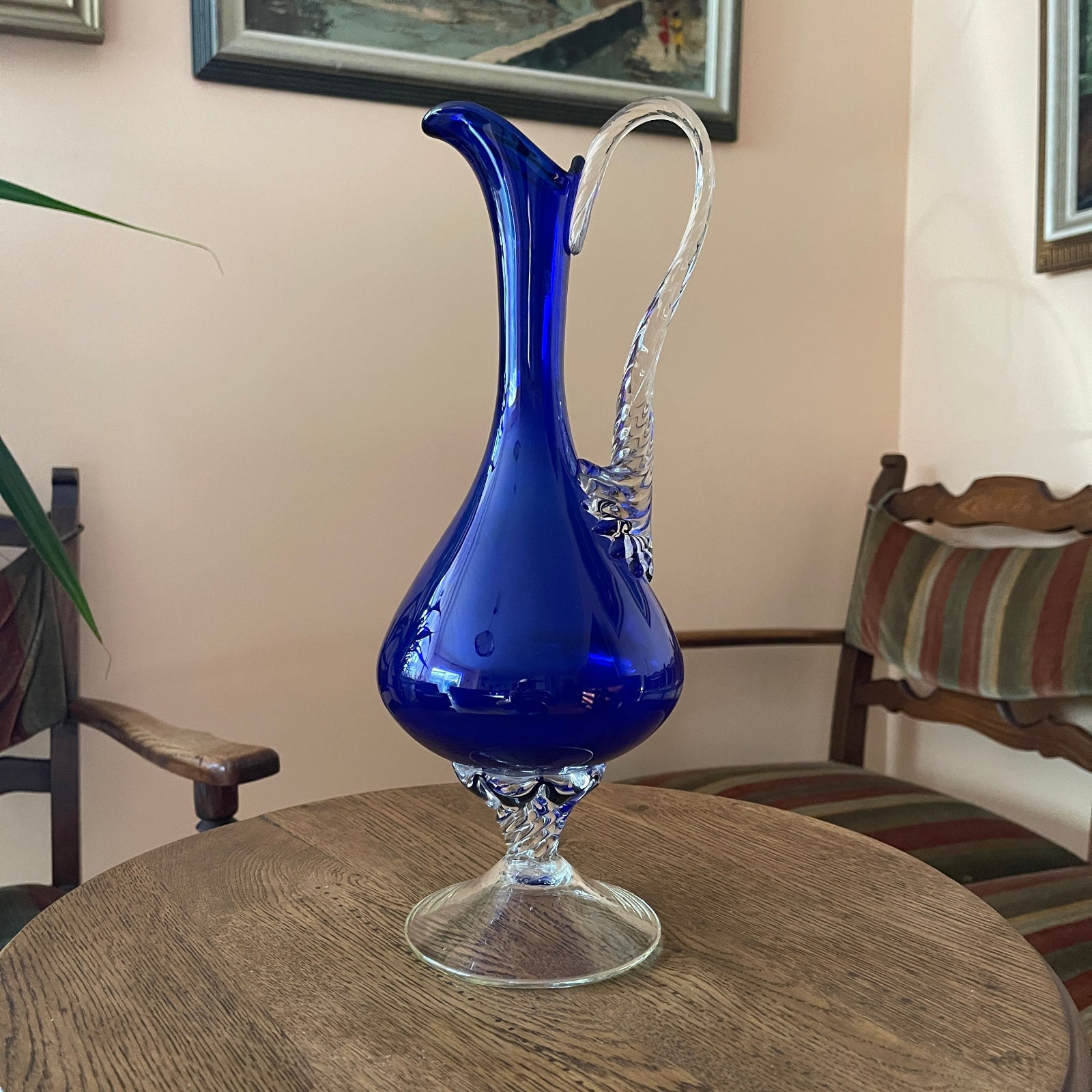 Blue glass pitcher on a wooden table with a chair and wall art in the background