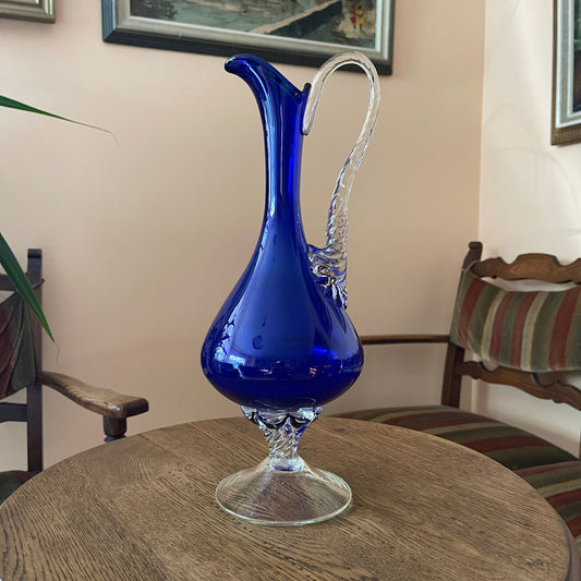 Blue glass pitcher on a wooden table with a chair and wall art in the background