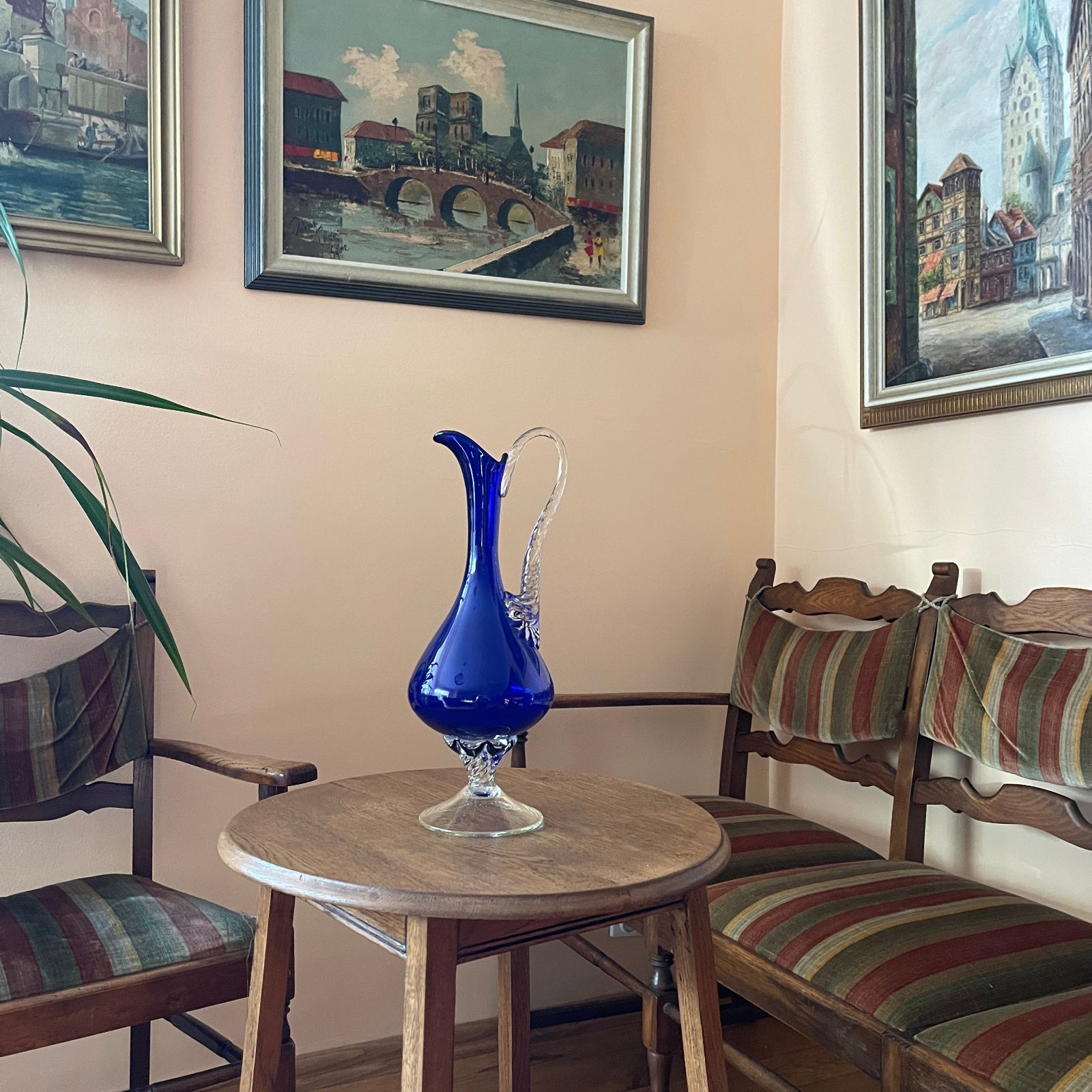 Blue glass pitcher on a wooden table with striped chairs and framed paintings in a room.