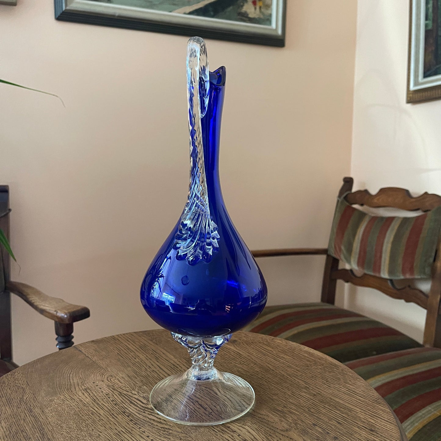 Blue glass vase on a wooden table with chairs and a wall painting in the background