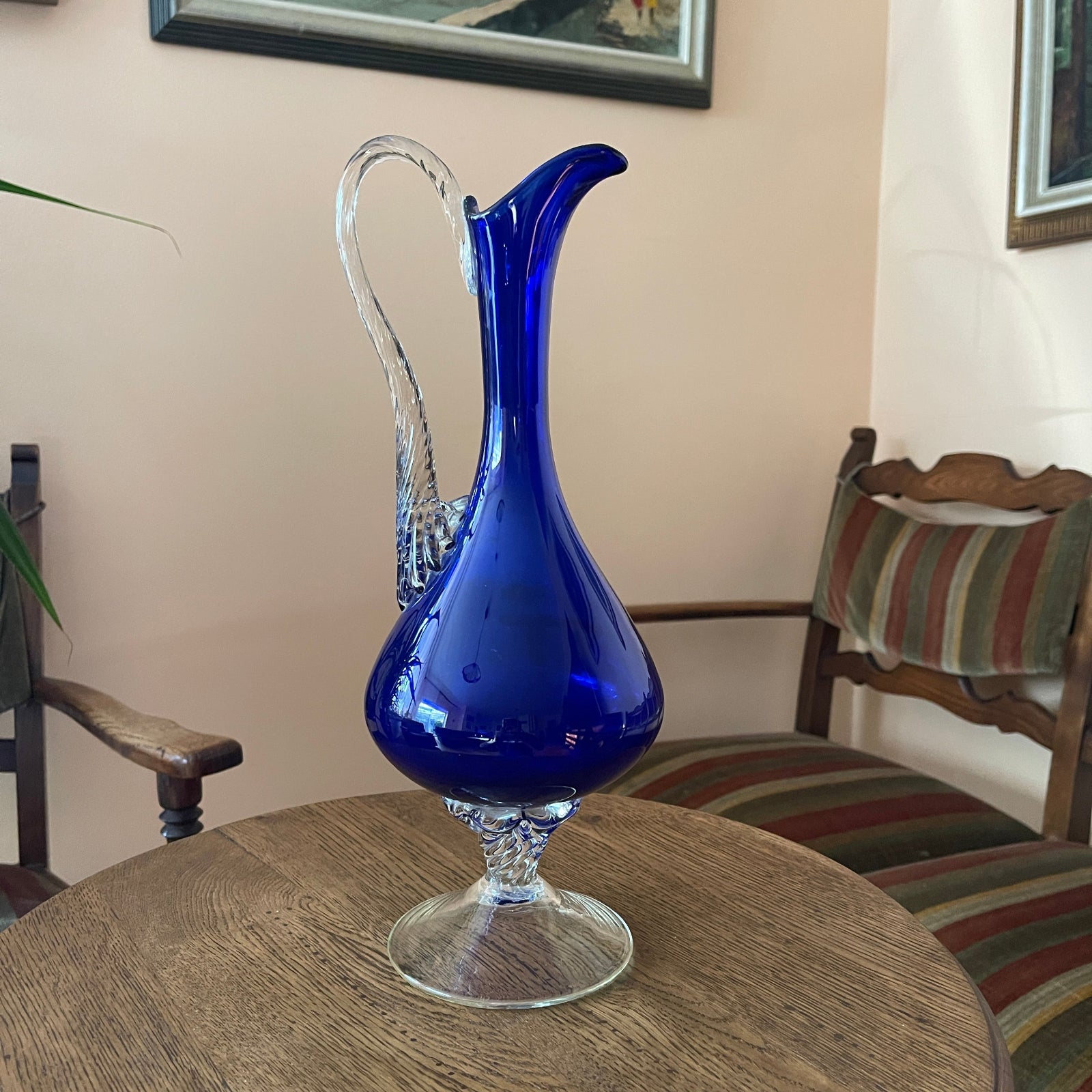 Blue glass pitcher on a wooden table with chairs and a wall painting in the background