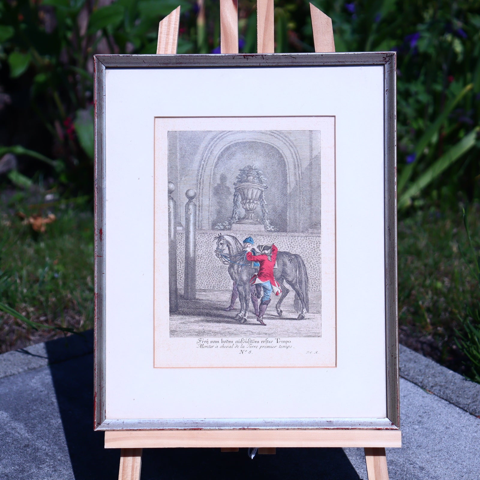 Framed artwork of two figures embracing a horse on an easel outdoors.
