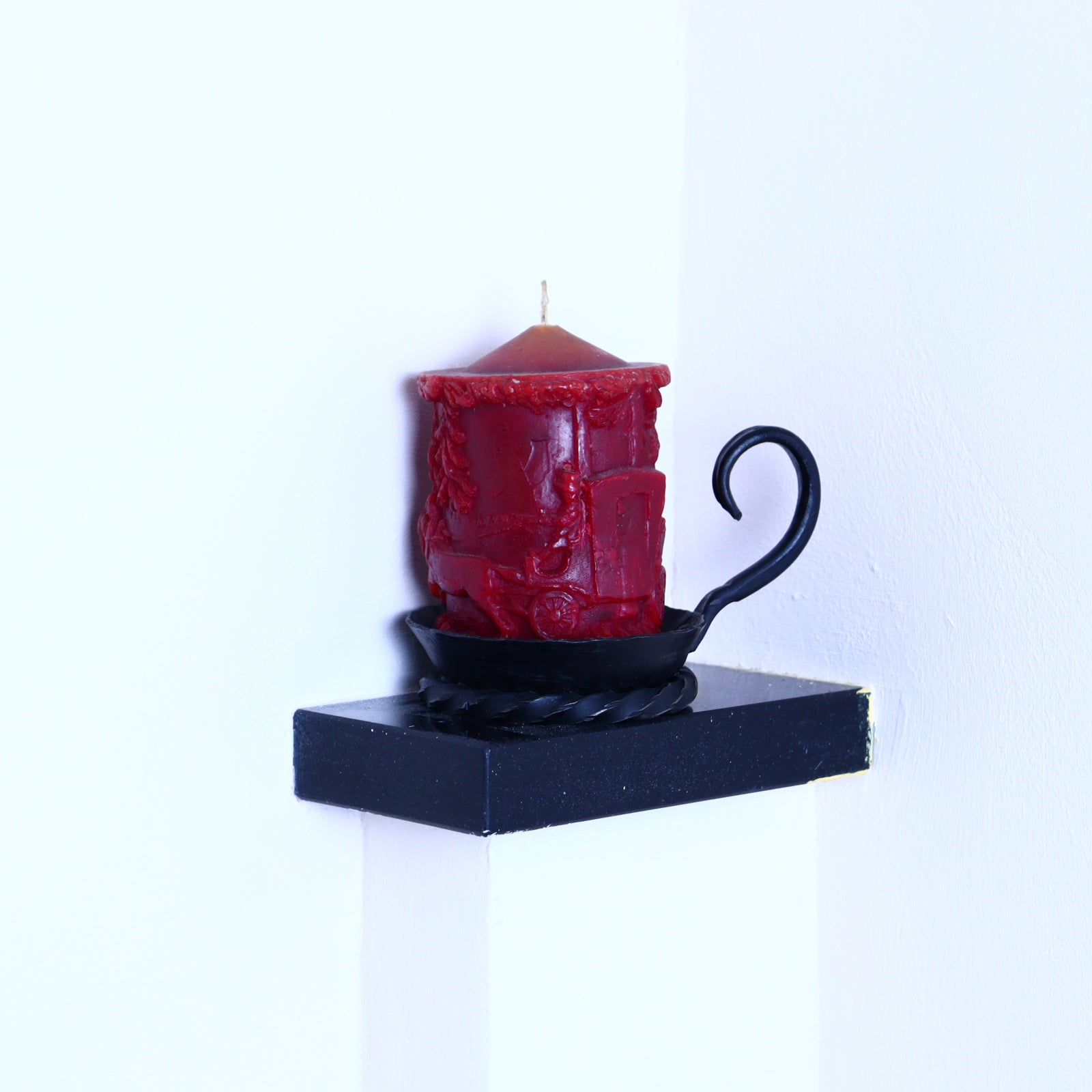 A red candle with a black lid and a twisted black handle, placed on a black stand against a white wall.
