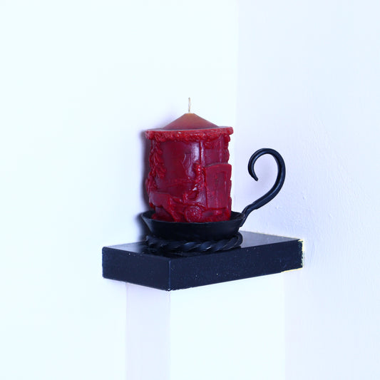 A red candle with a black lid and a twisted black handle, placed on a black stand against a white wall.