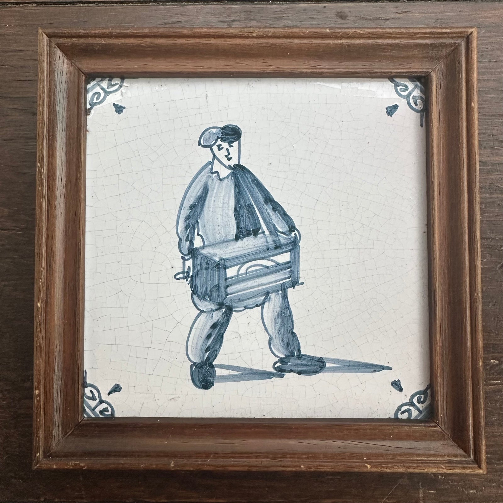 Framed tile with a blue and white illustration of a man carrying a crate on a wooden surface