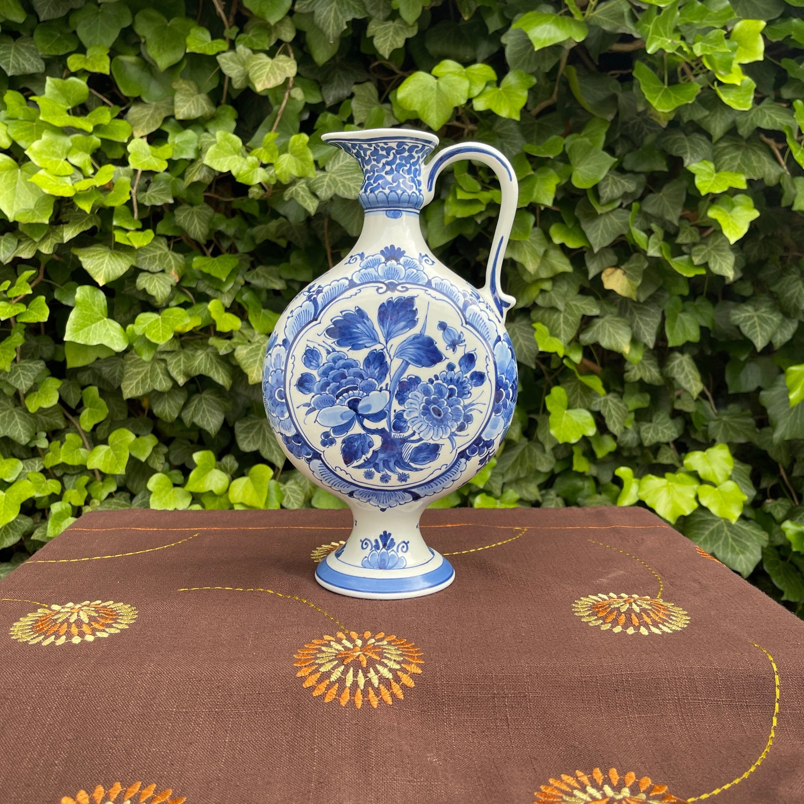 Blue and white floral ceramic pitcher on a brown tablecloth with a green leafy background