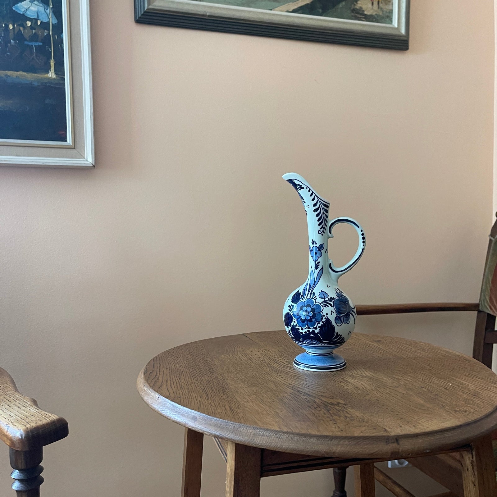 Blue and white ceramic pitcher on a wooden table against a beige wall with framed pictures.