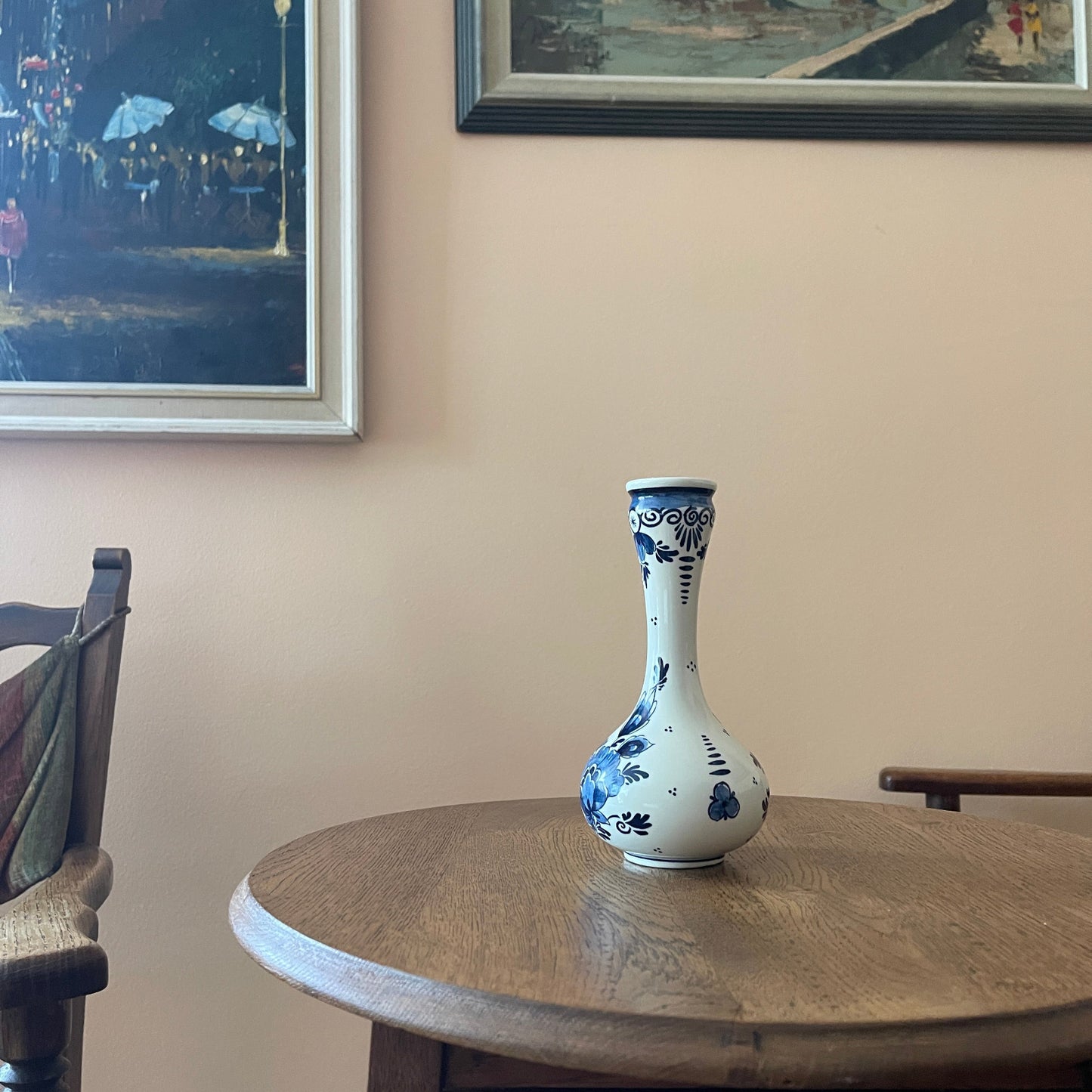 White vase with blue patterns on a wooden table against a beige wall with framed artwork.