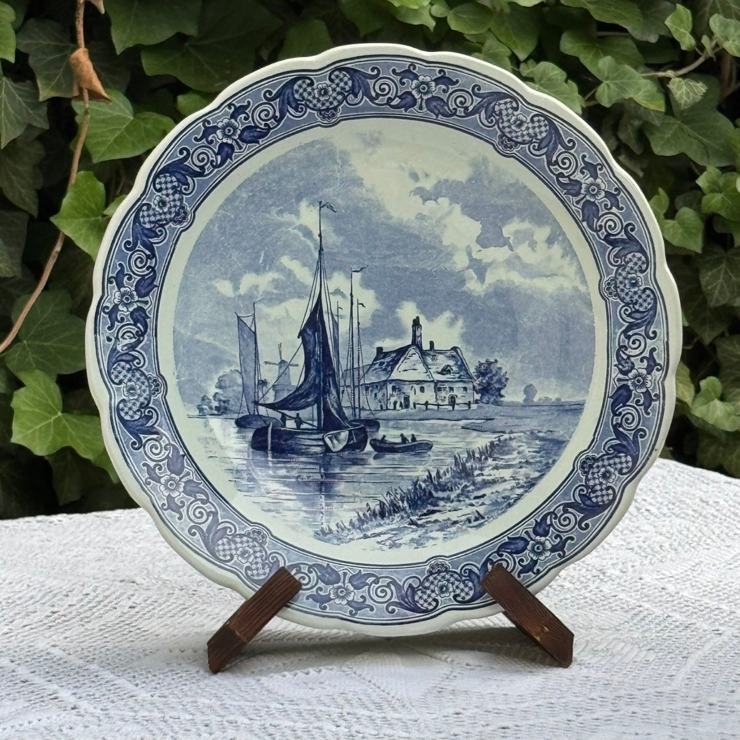 Decorative plate with blue and white design of a sailboat and house, placed on a stand against a green leafy background.