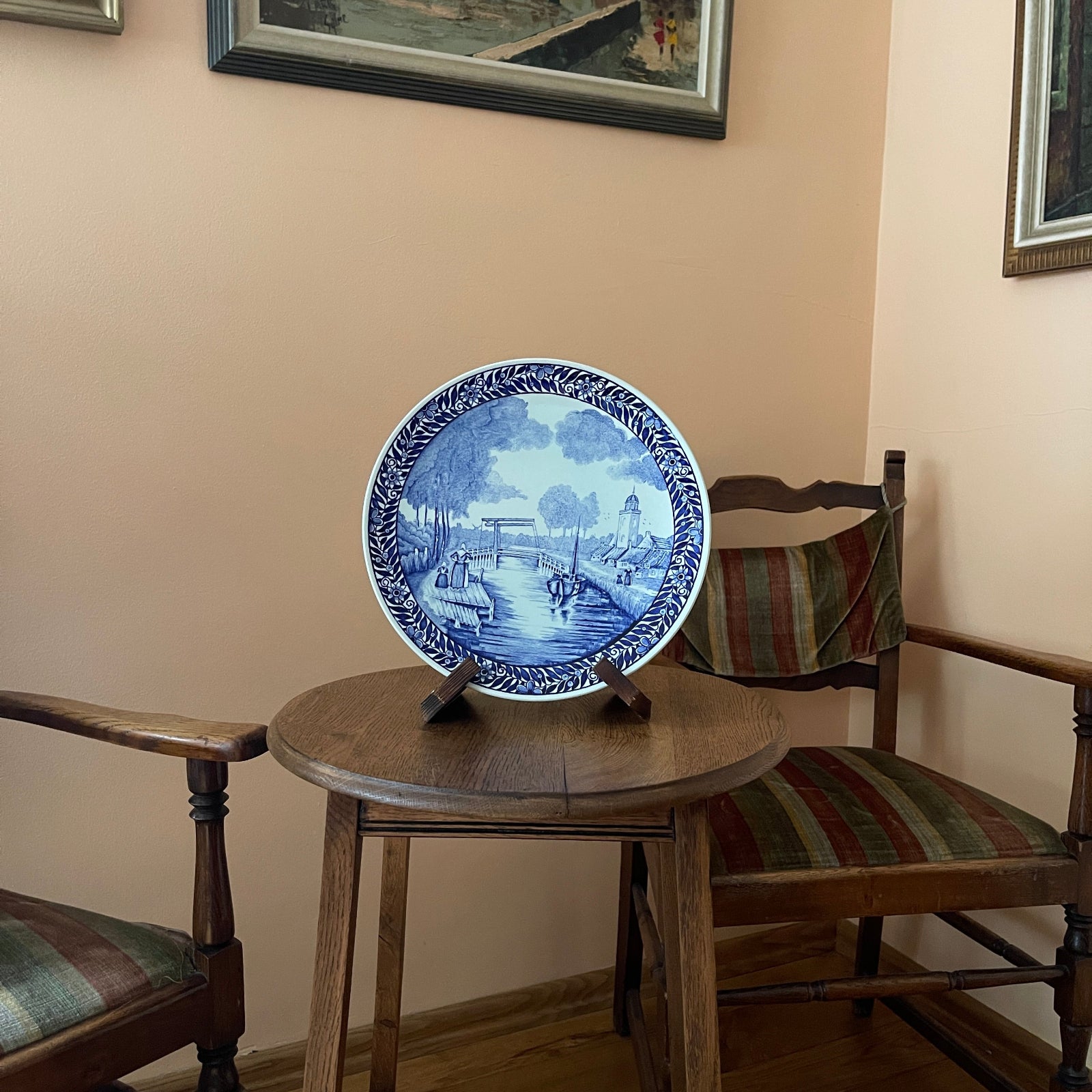Blue and white decorative plate on a wooden table with chairs and framed pictures in the background.