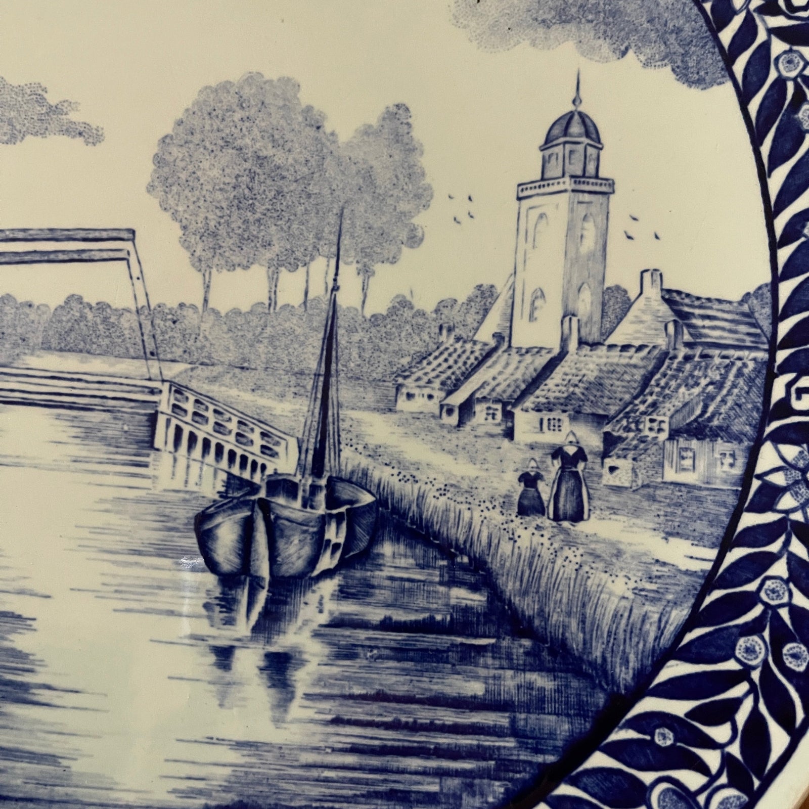 Decorative tile with a blue and white scene of a village by a river with a boat and a clock tower.