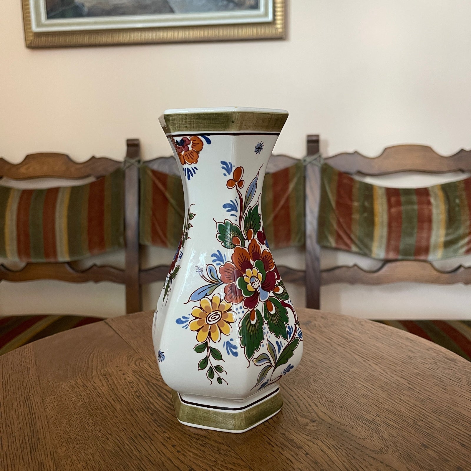 Decorative vase with floral patterns on a wooden table in a room with chairs and a painting.