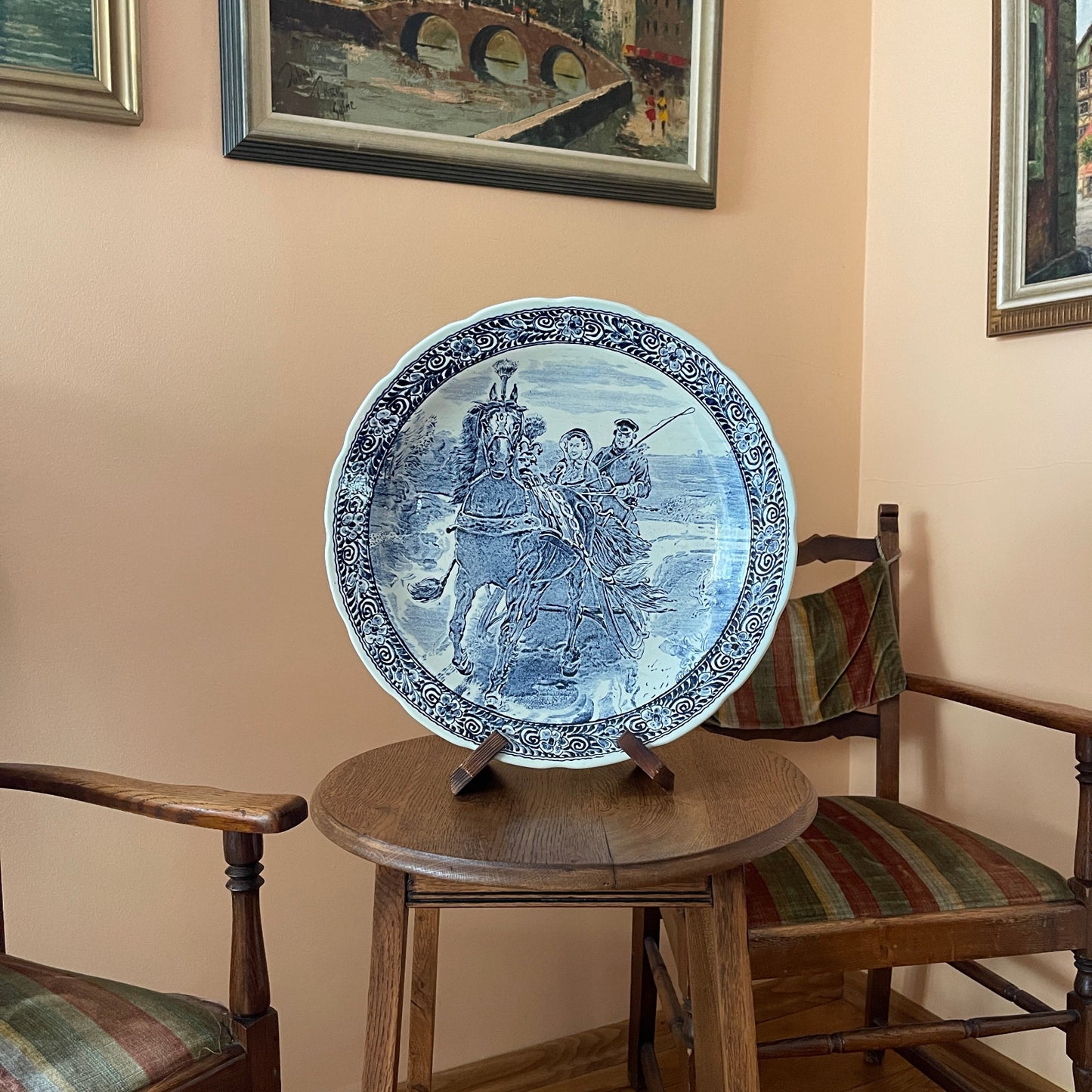Decorative blue and white plate on a wooden table with chairs and framed pictures in the background.