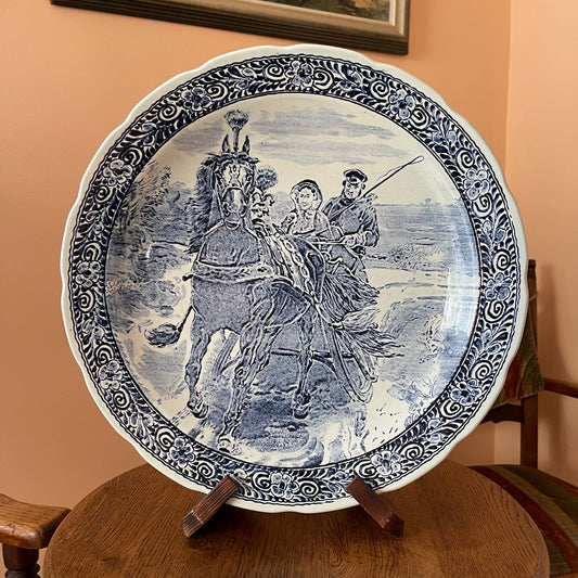 Decorative plate with a blue and white design of a scene with people and a horse on a wooden stand.